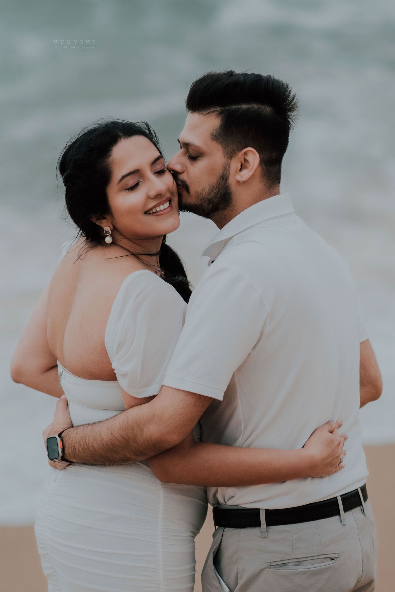 Couple sharing a tender kiss on a sandy beach, woman in white dress smiling in a romantic embrace.