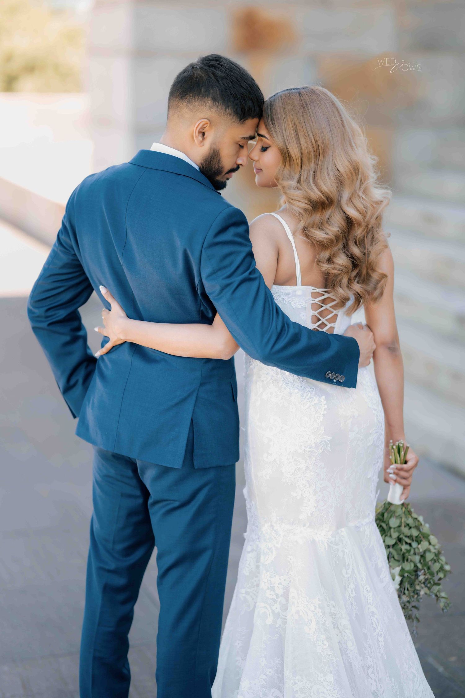 Bride and groom embrace tenderly from behind in elegant wedding attire outdoors.