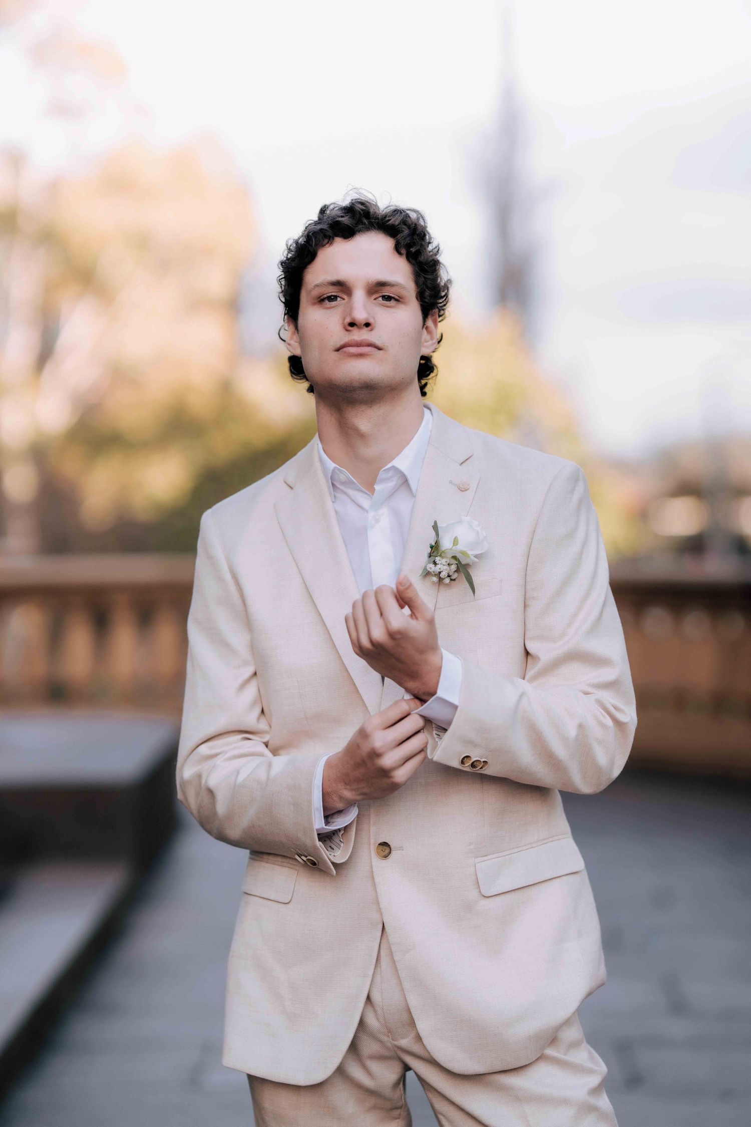 Groom in beige wedding suit adjusting cufflinks with white floral boutonniere outdoors.