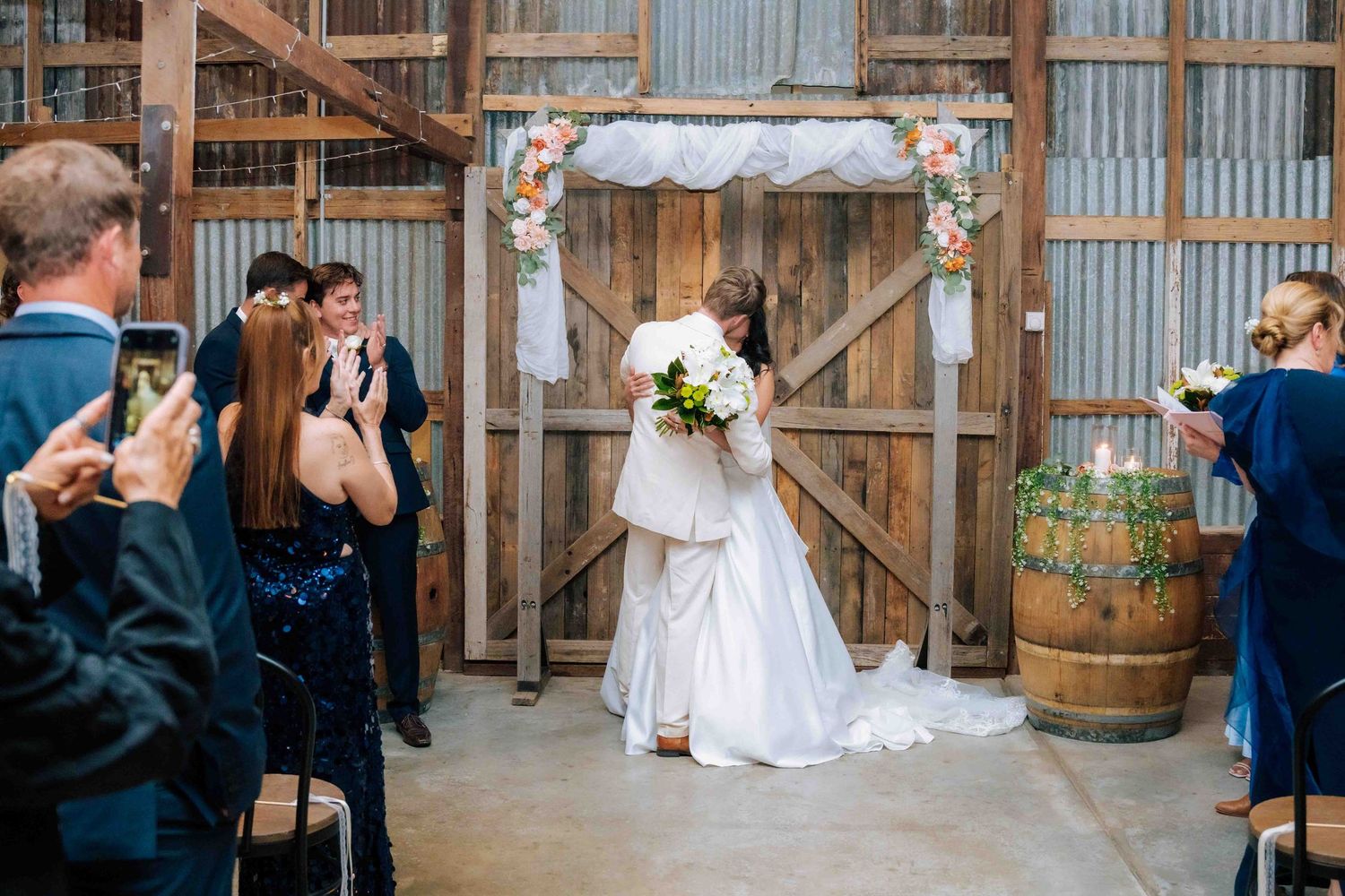 Bride and groom share first kiss at rustic barn wedding ceremony with floral arch, guests applauding and taking photos.