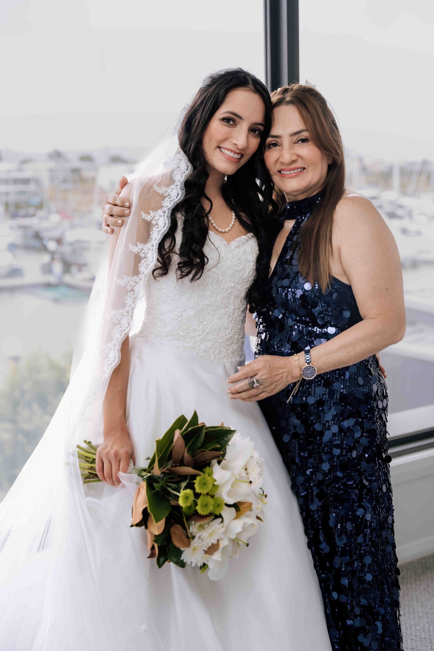 Smiling bride in lace wedding gown holds bouquet posing with mother in navy sequin dress.