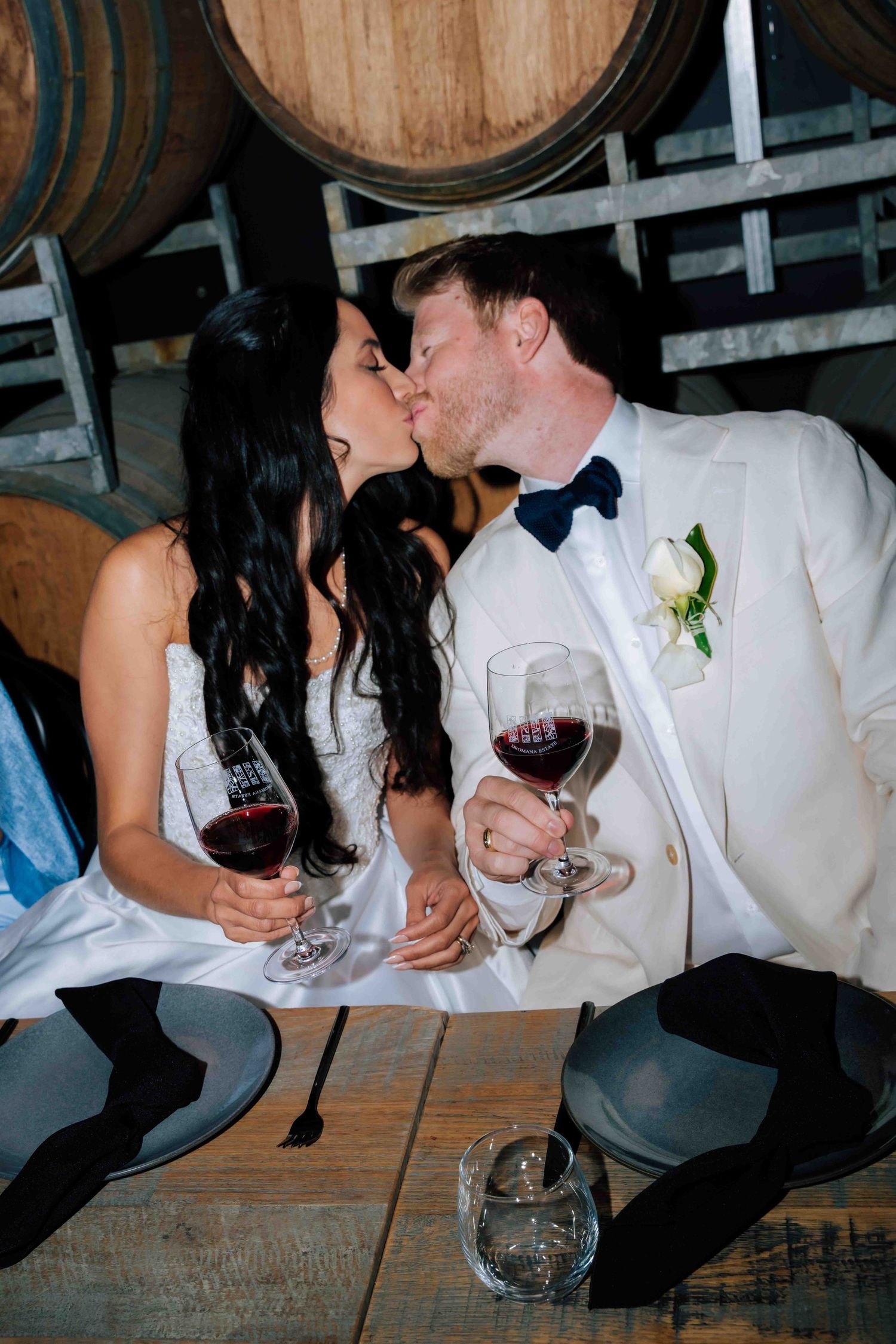 Bride and groom kiss while holding red wine glasses seated among oak barrels at winery wedding reception.