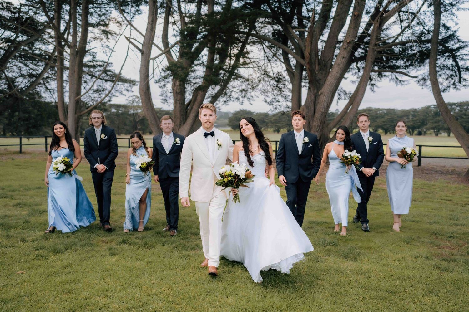 Wedding party walks outdoors on green grass with trees, bride in white gown and groom in cream suit leading bridesmaids and groomsmen.