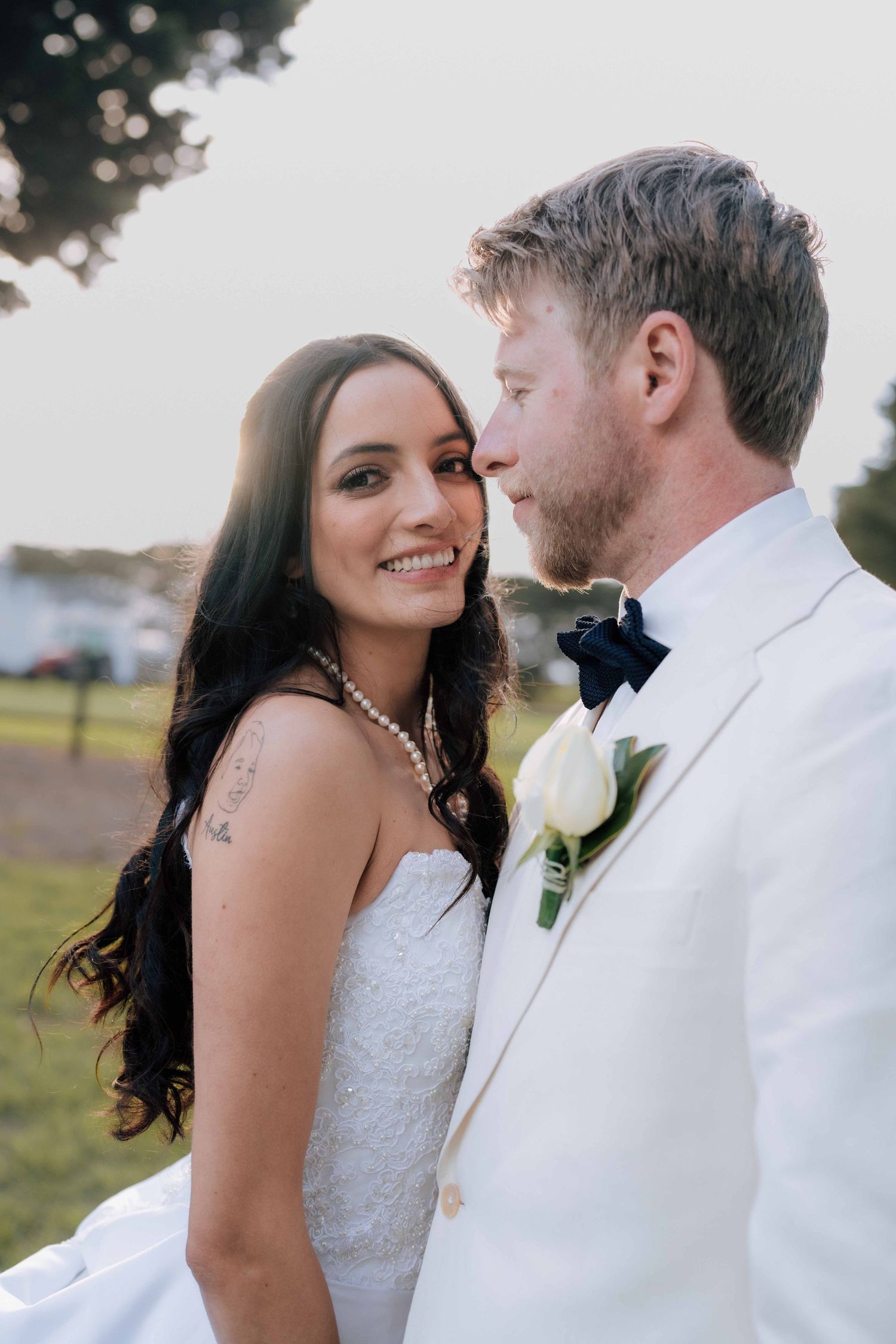 Smiling bride in white lace gown wearing pearl necklace poses closely with groom in white suit at outdoor wedding.
