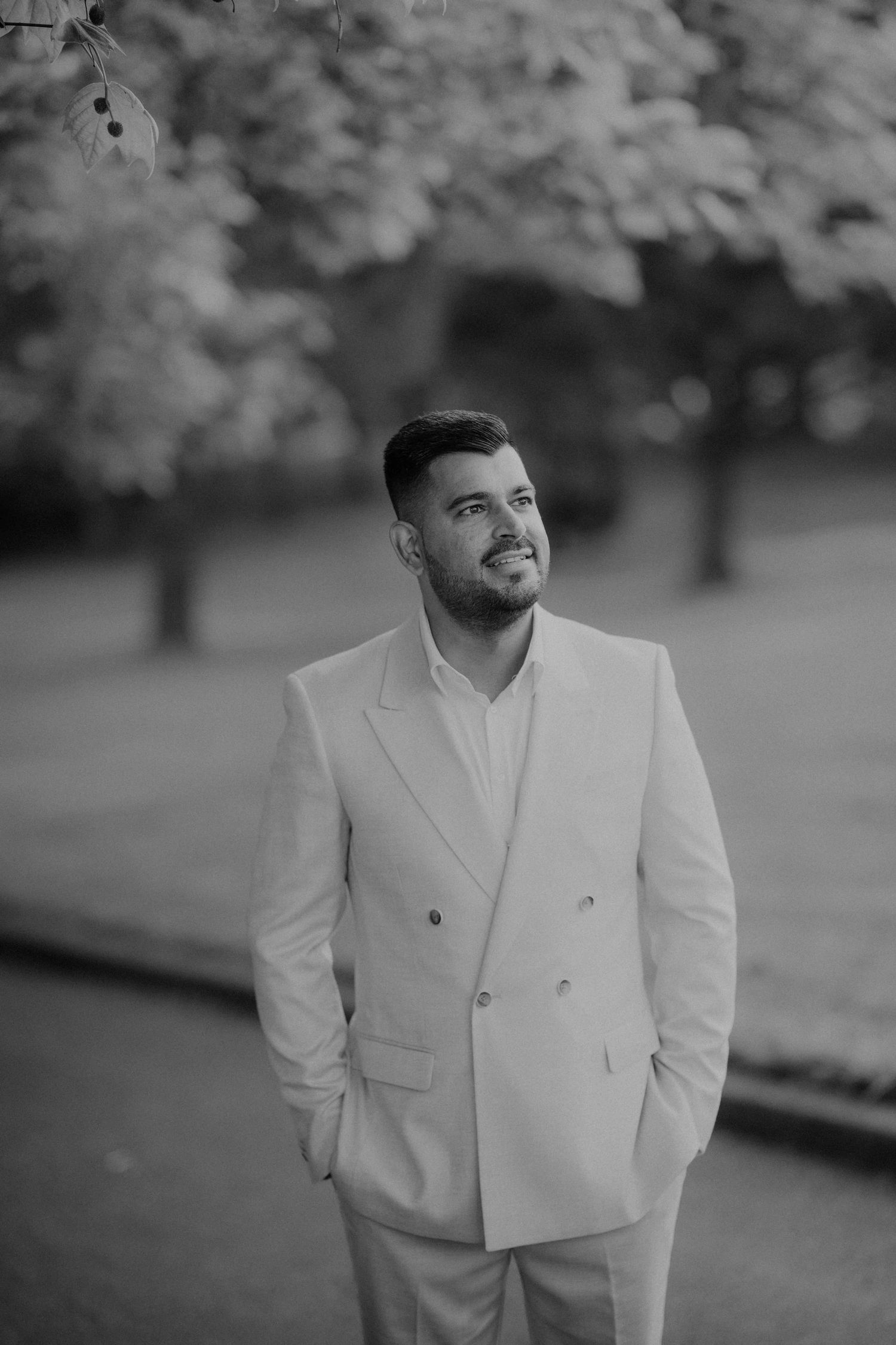 Black and white portrait of a groom in a double-breasted white suit standing in a park.