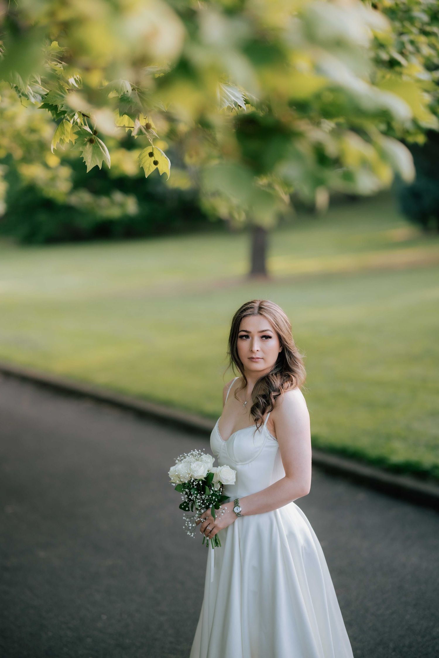 Elegant bride in a sweetheart neckline gown holding a white bouquet standing on a tree-lined park pathway.