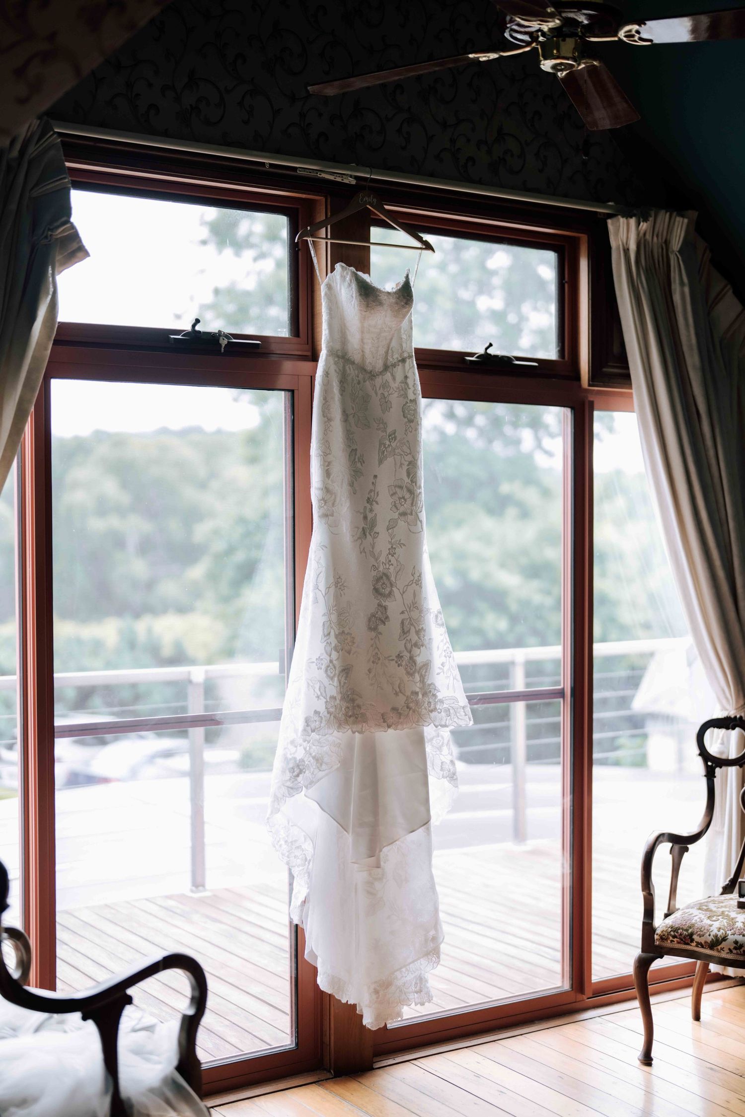 A lace wedding dress hanging in front of large windows with a lush green garden view outside.