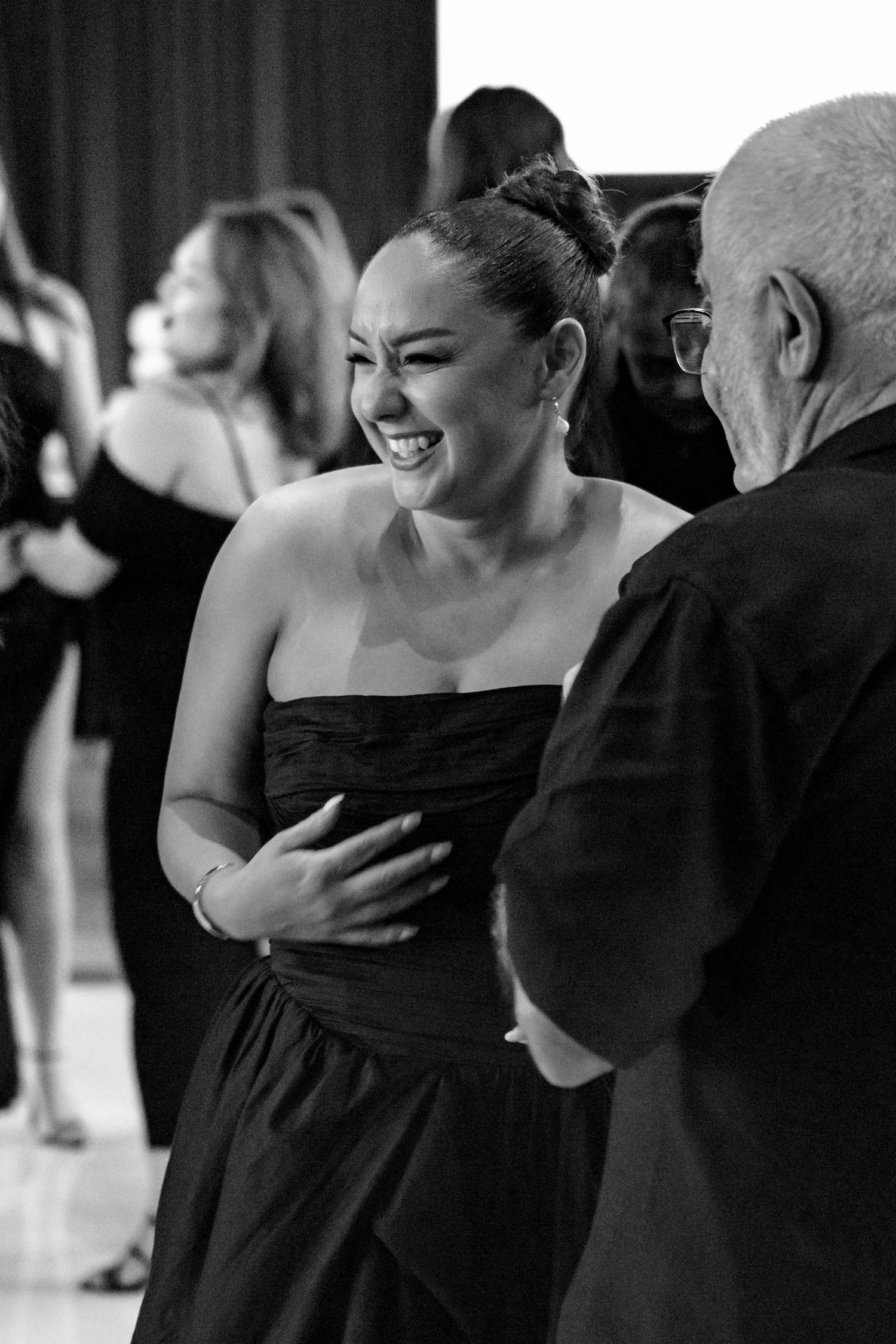 A woman in a black strapless dress laughing joyfully while socializing at a wedding reception.