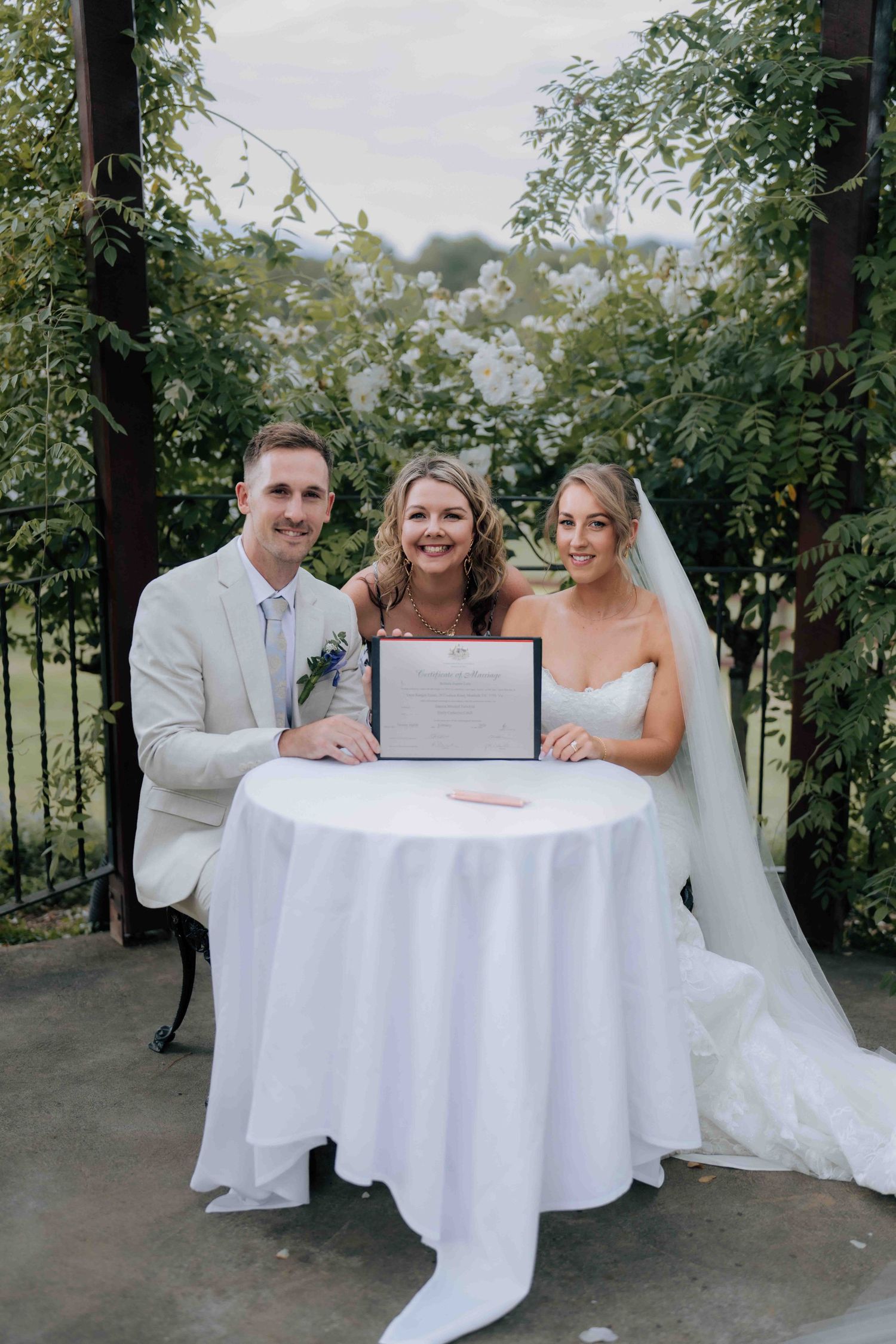 Bride, groom and celebrant posing with marriage certificate at outdoor garden wedding ceremony.