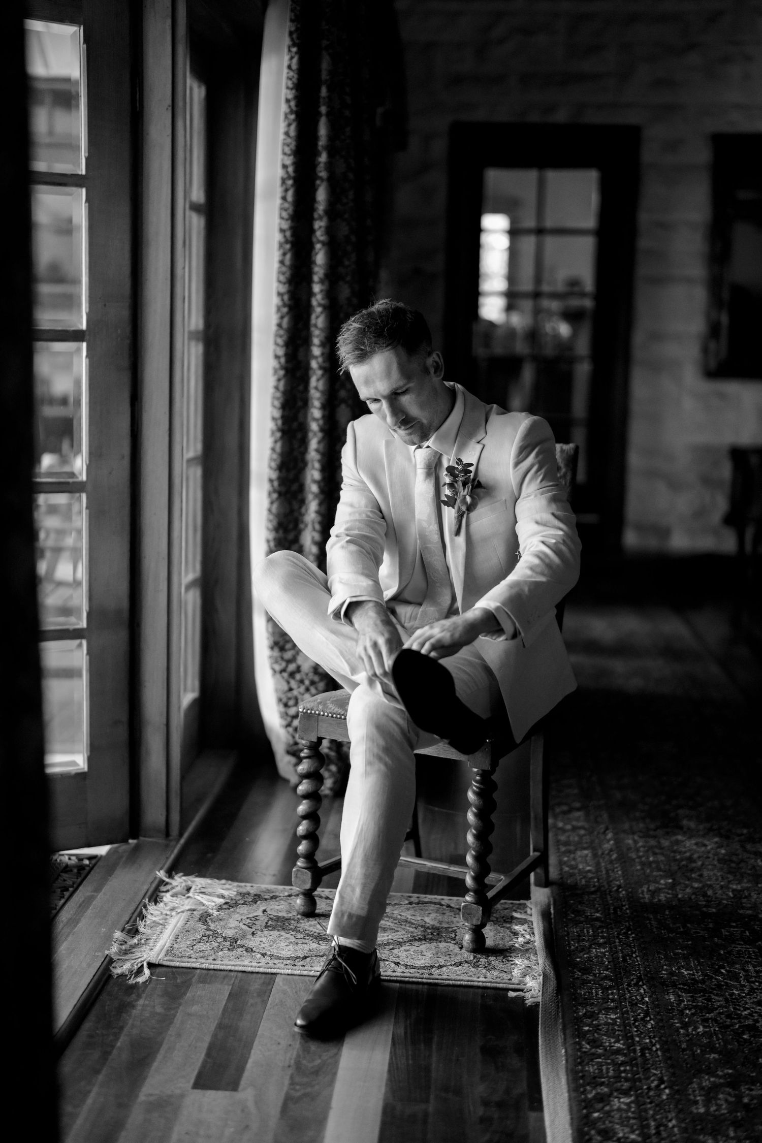 Groom in white suit puts on shoes while seated by window in black and white wedding photo.
