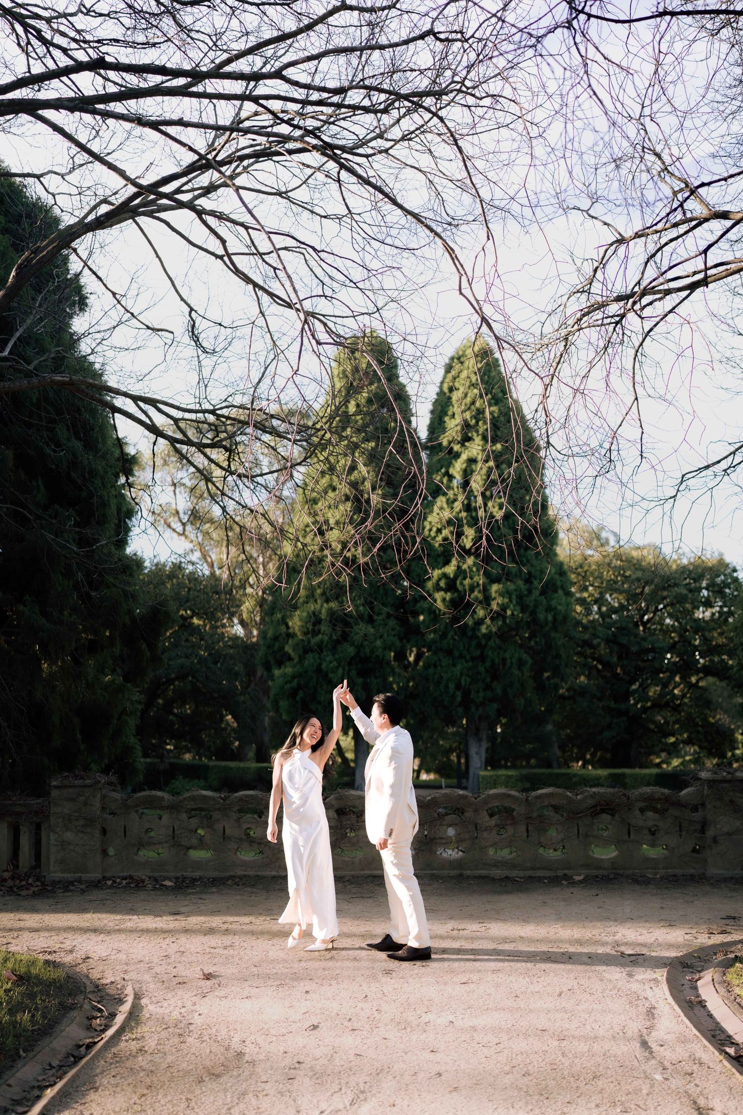 Couple in white outfits dancing together in a scenic park with tall cypress trees and bare branches overhead.