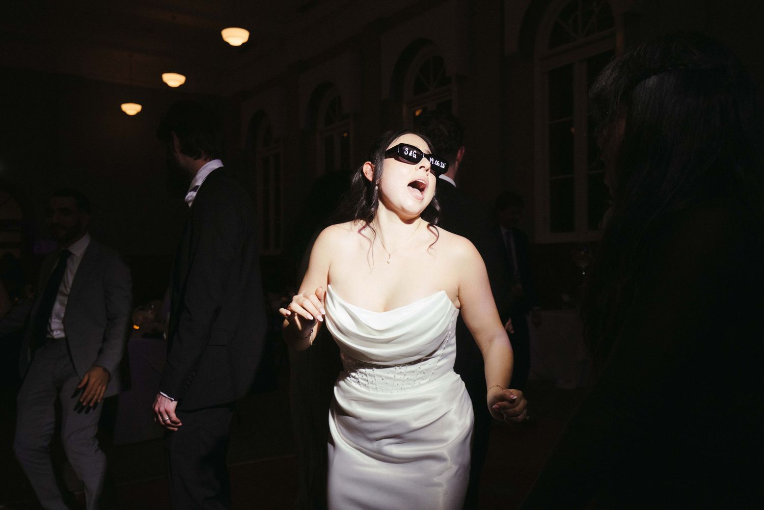 Bride wearing sunglasses dancing energetically at dark wedding reception venue.