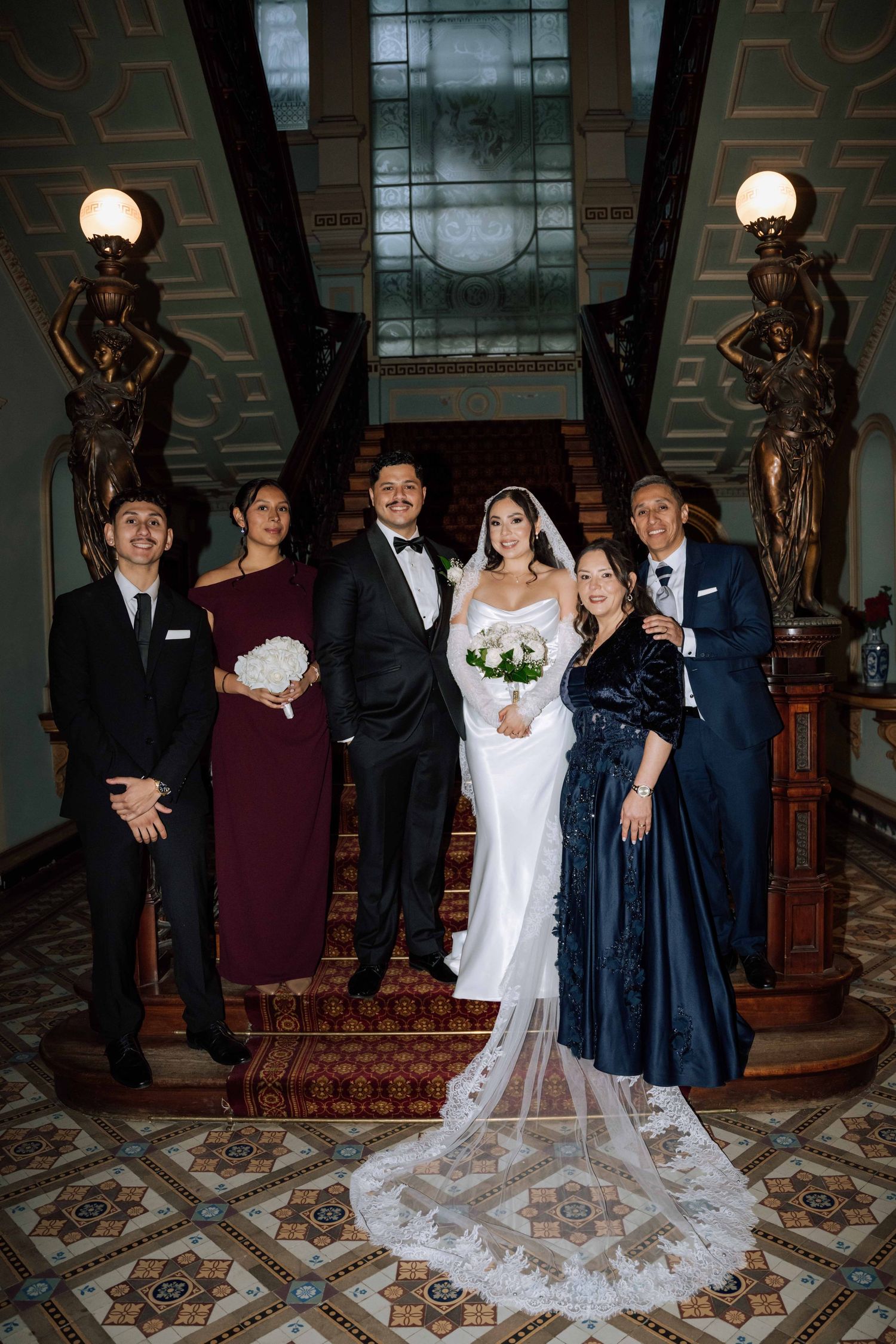 Wedding party poses on ornate grand staircase inside historic venue with decorative statues and tiles.