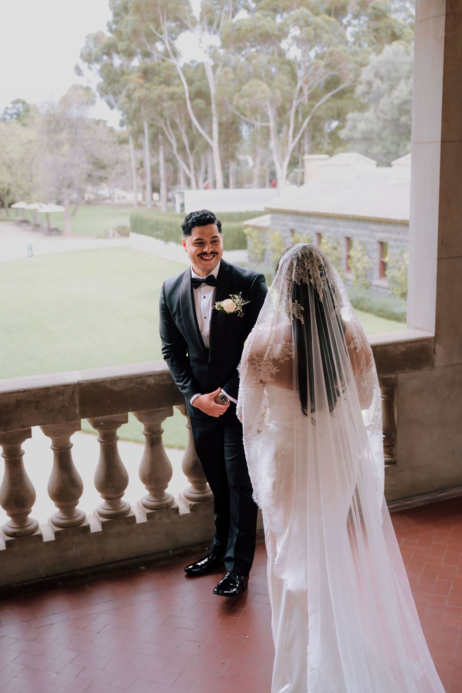 Groom in black tuxedo smiling at bride in lace wedding dress and veil on elegant balcony.