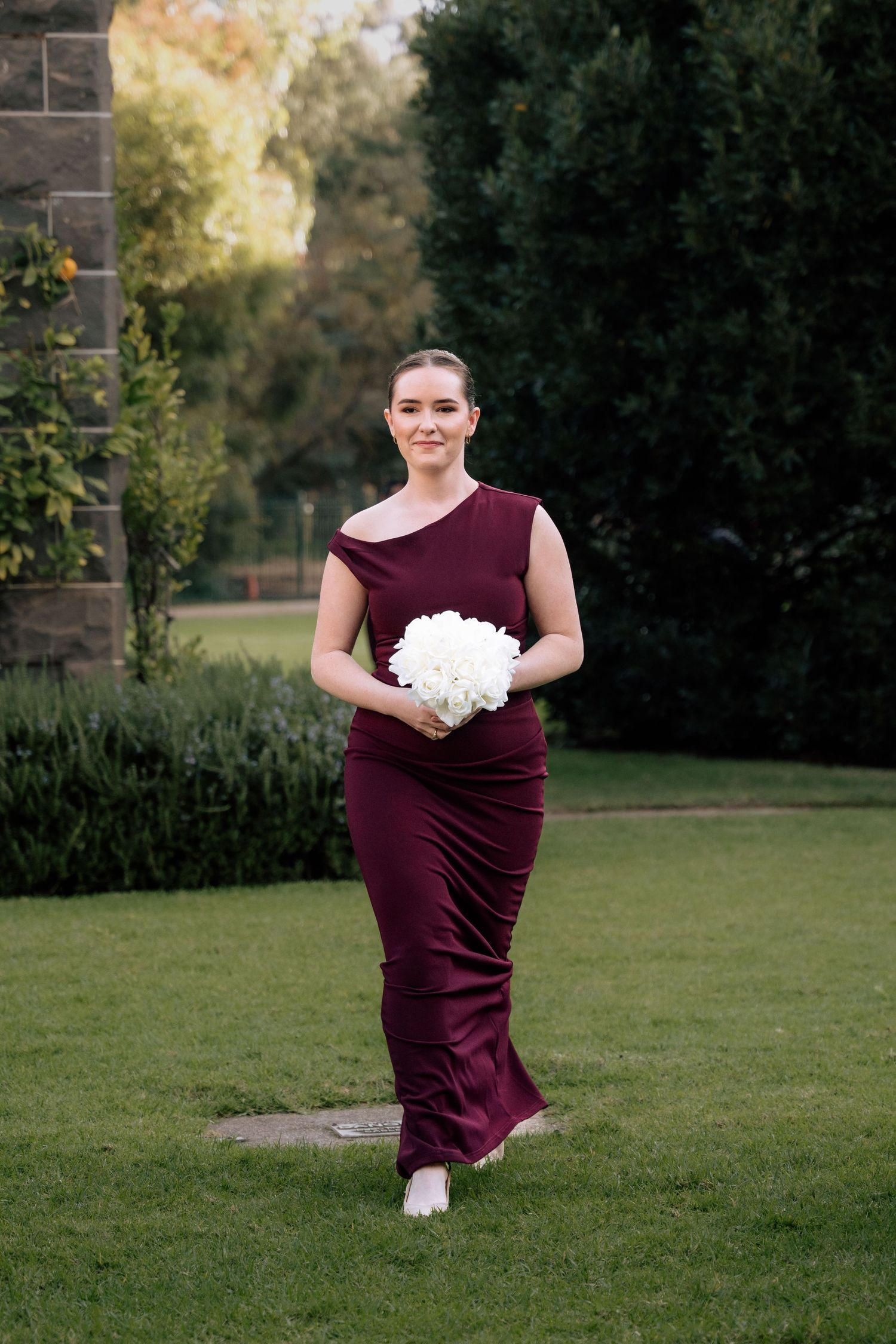 Bridesmaid in burgundy one-shoulder gown walking across garden lawn holding white hydrangea bouquet.