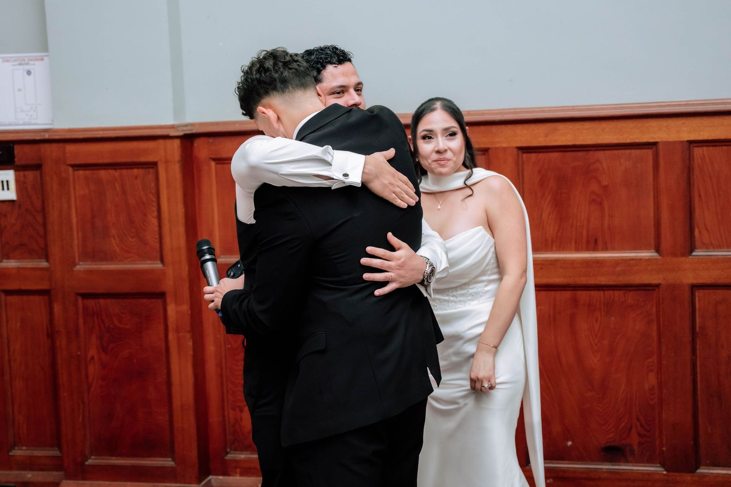 Groom shares emotional hug with guest while bride in white satin gown watches at wedding reception.