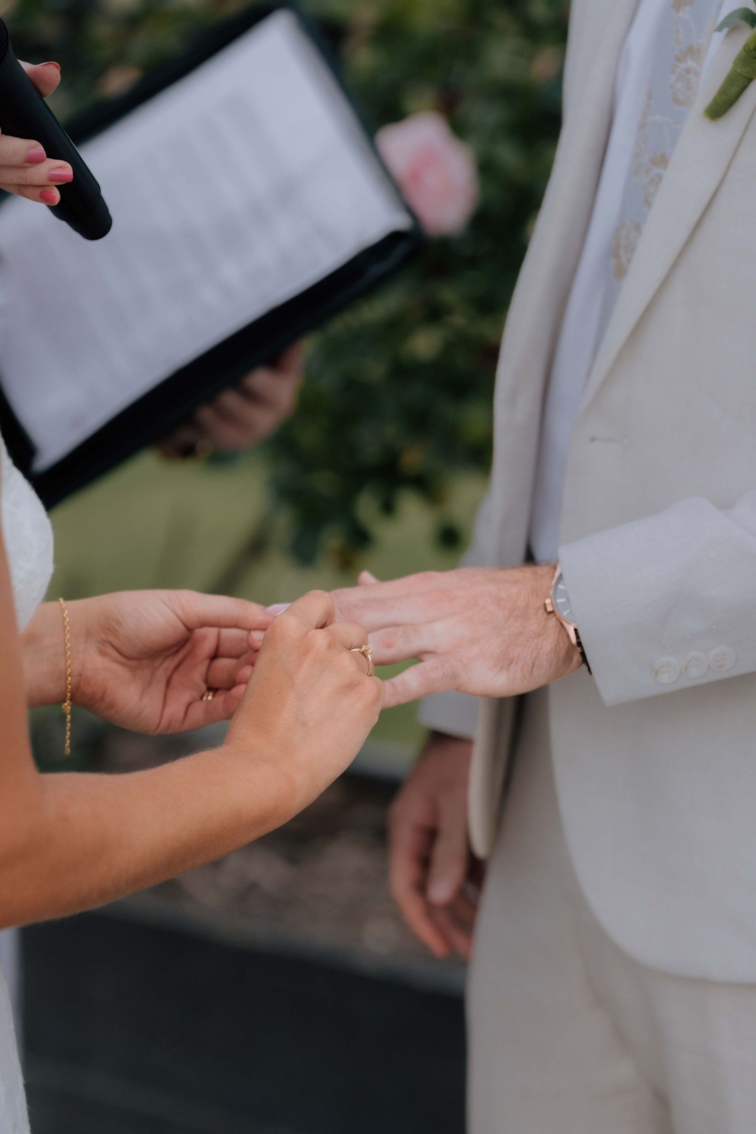 Bride sliding wedding ring onto groom's finger during outdoor wedding ceremony with officiant holding black folder.