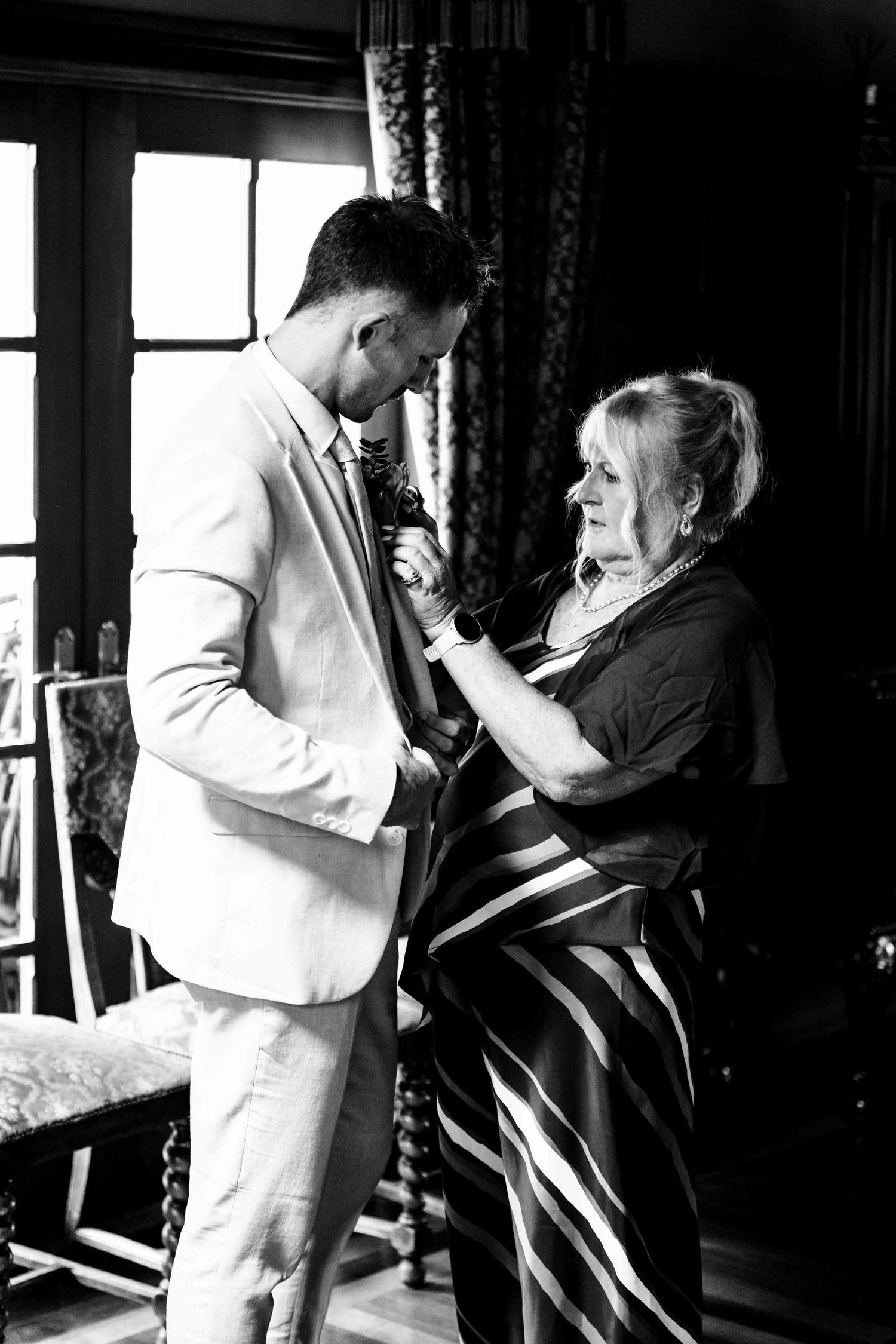 Mother helping groom pin boutonniere on suit lapel in black and white wedding preparation photo.