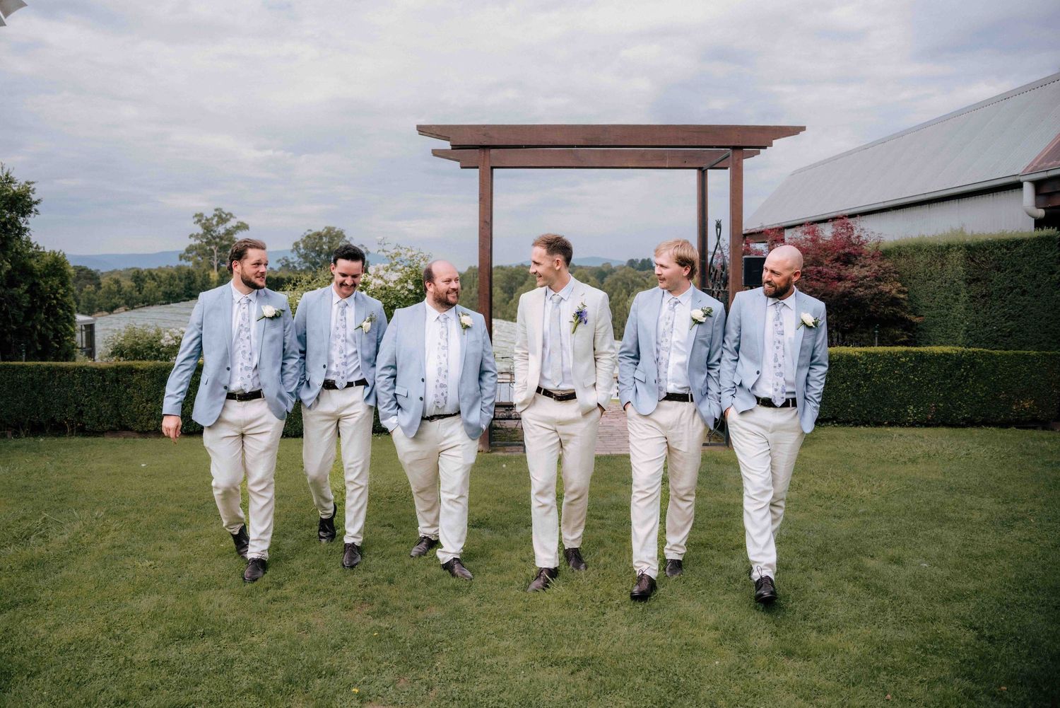Six groomsmen in matching blue jackets and khaki pants walking casually across green lawn.