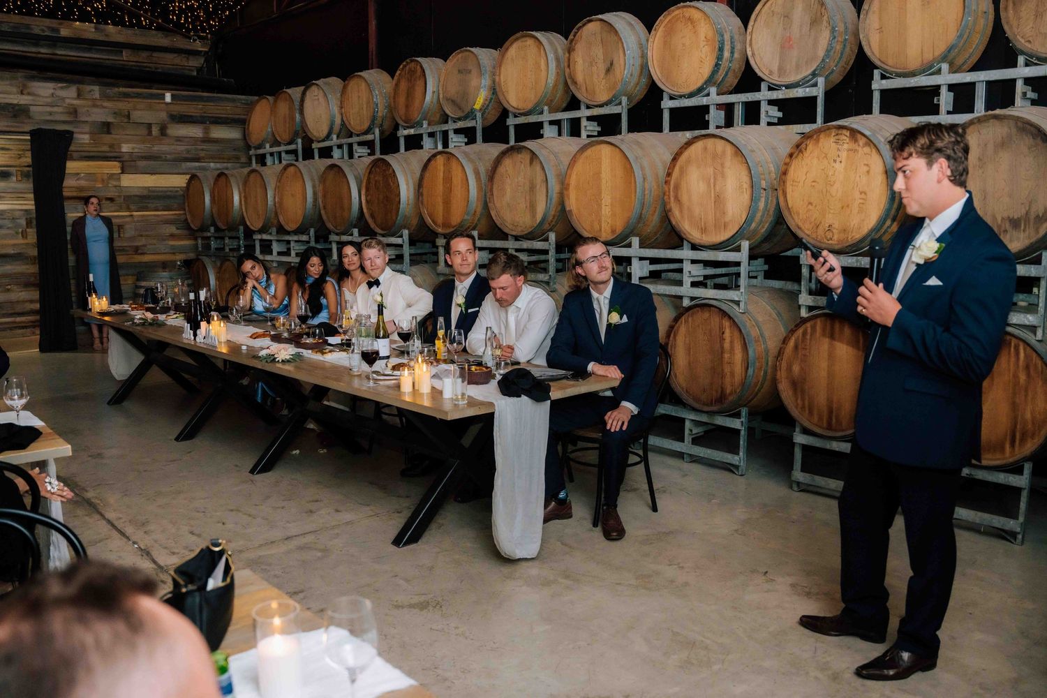 Groomsman delivers a speech to guests seated at a long table surrounded by oak wine barrels at a winery wedding.