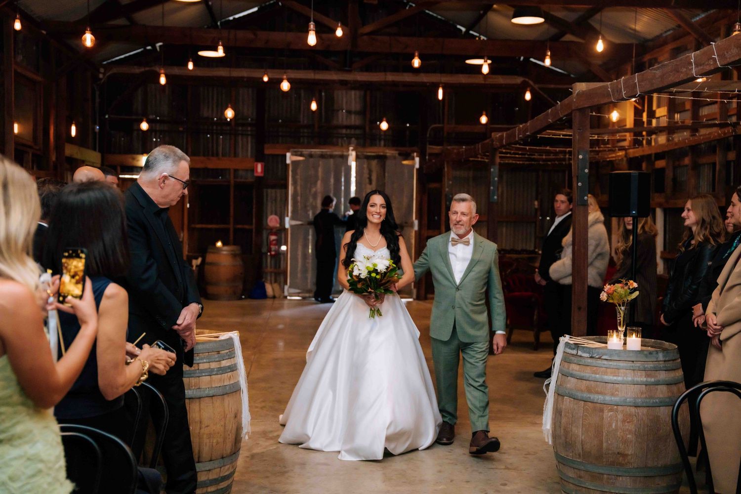 Bride in white ball gown walking down aisle in rustic barn venue with string lights.