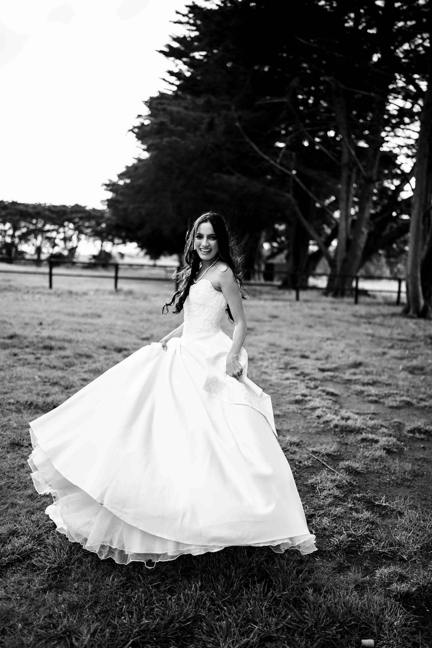 Black and white photo of smiling bride twirling her elegant ball gown on a grassy outdoor setting.