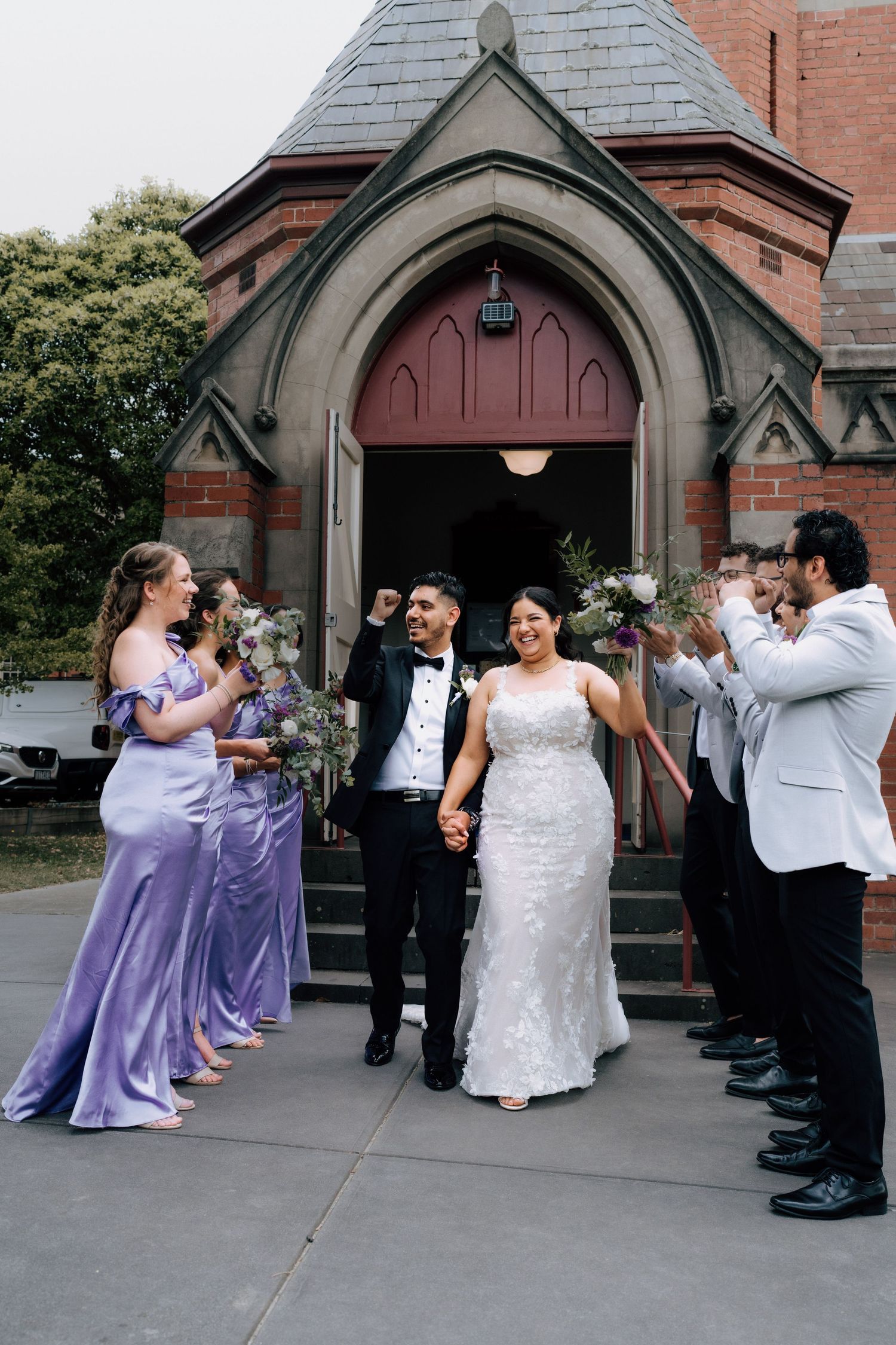 Bride and groom exit church as wedding party in lavender dresses and white suits celebrate.