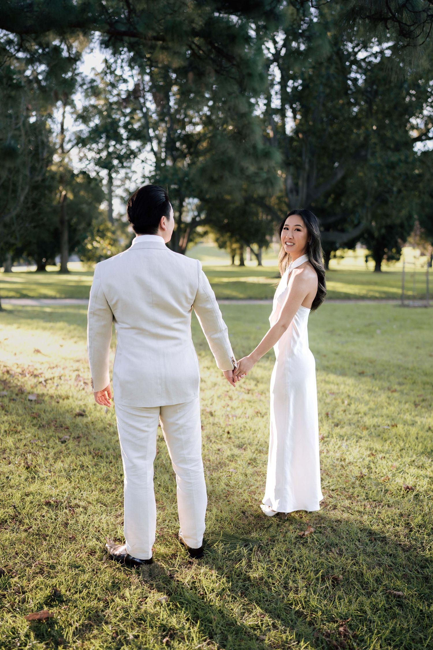 A smiling bride in a white halter jumpsuit holds hands with a groom in a cream suit in a sunlit park.