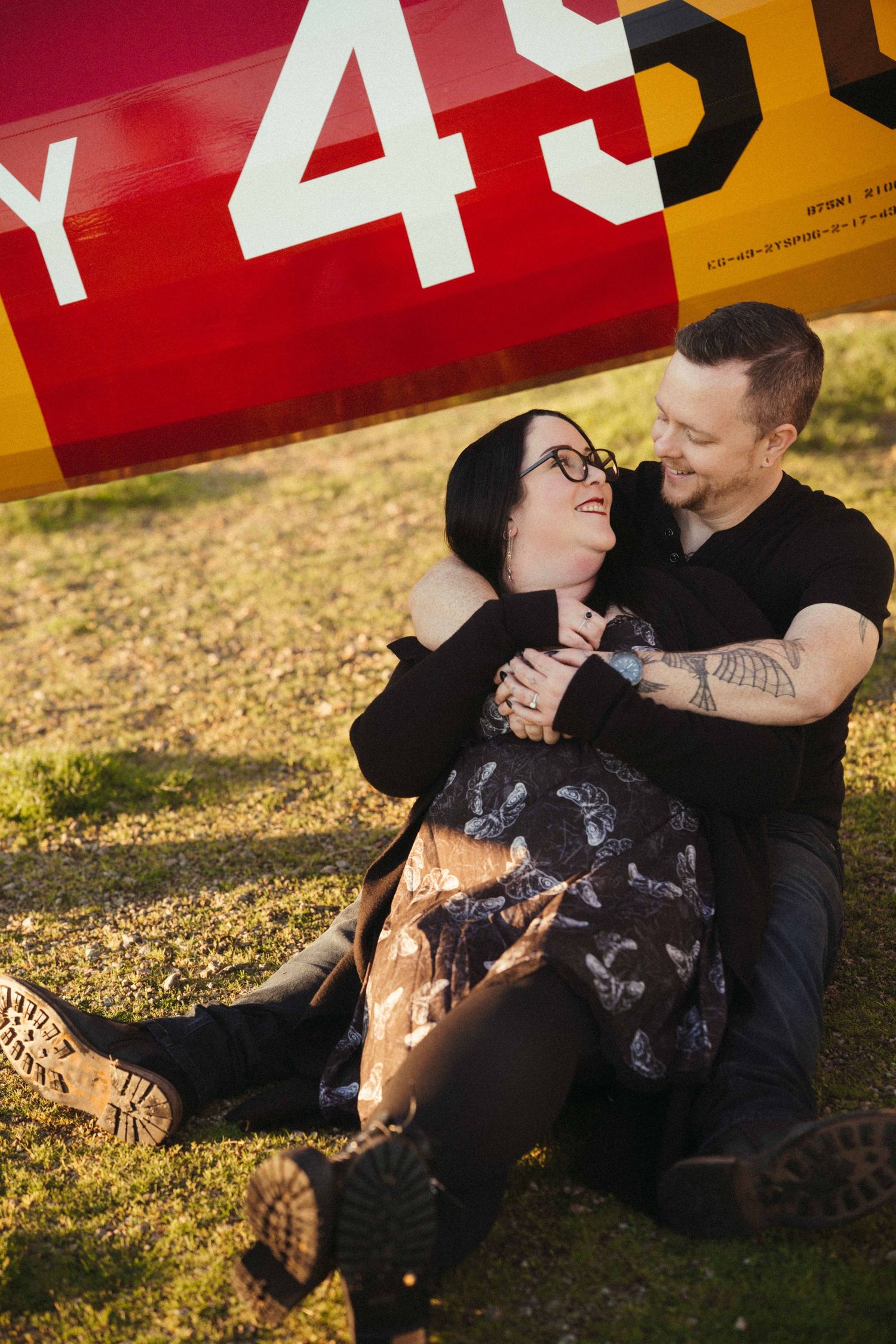 Couple sitting together on grass laughing in front of vintage yellow and red airplane outdoors.