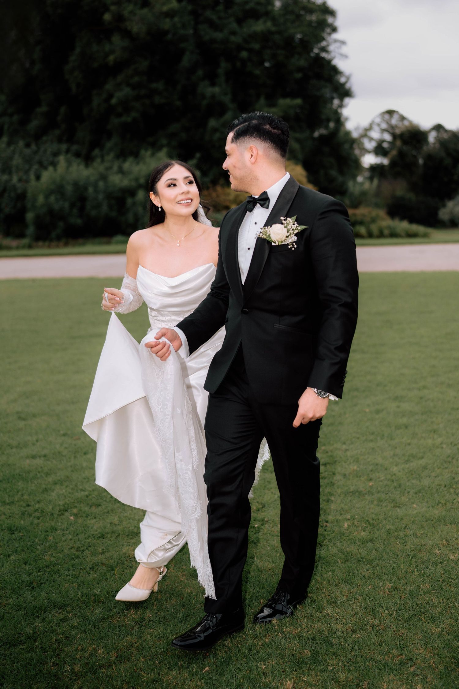 Bride in white off-shoulder gown and groom in black tuxedo walk hand-in-hand across a lush green lawn.