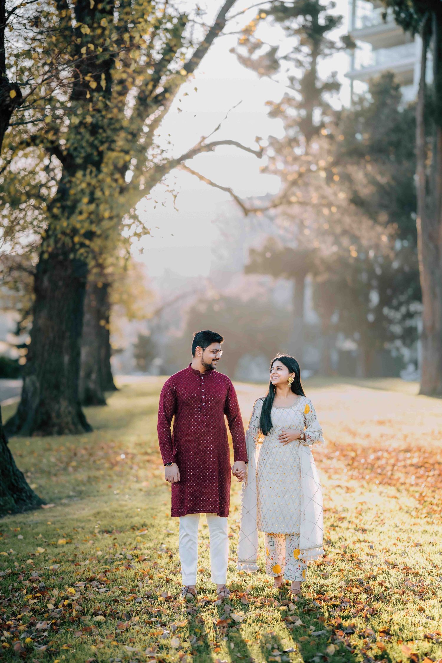 Indian couple in traditional attire holding hands walking through a sunlit park with golden autumn leaves.