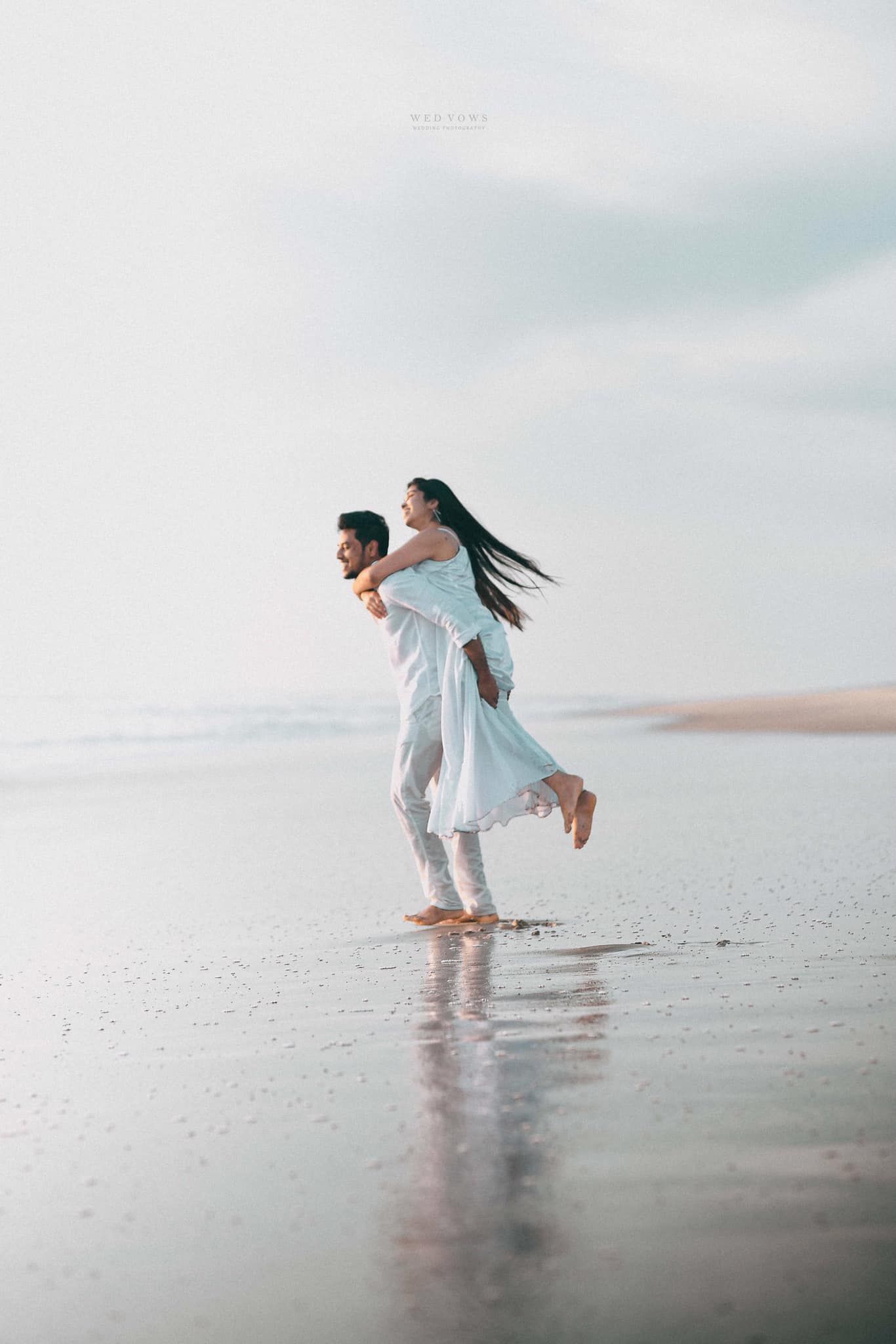 Man giving woman a piggyback ride on reflective wet beach shoreline, both dressed in white outfits.