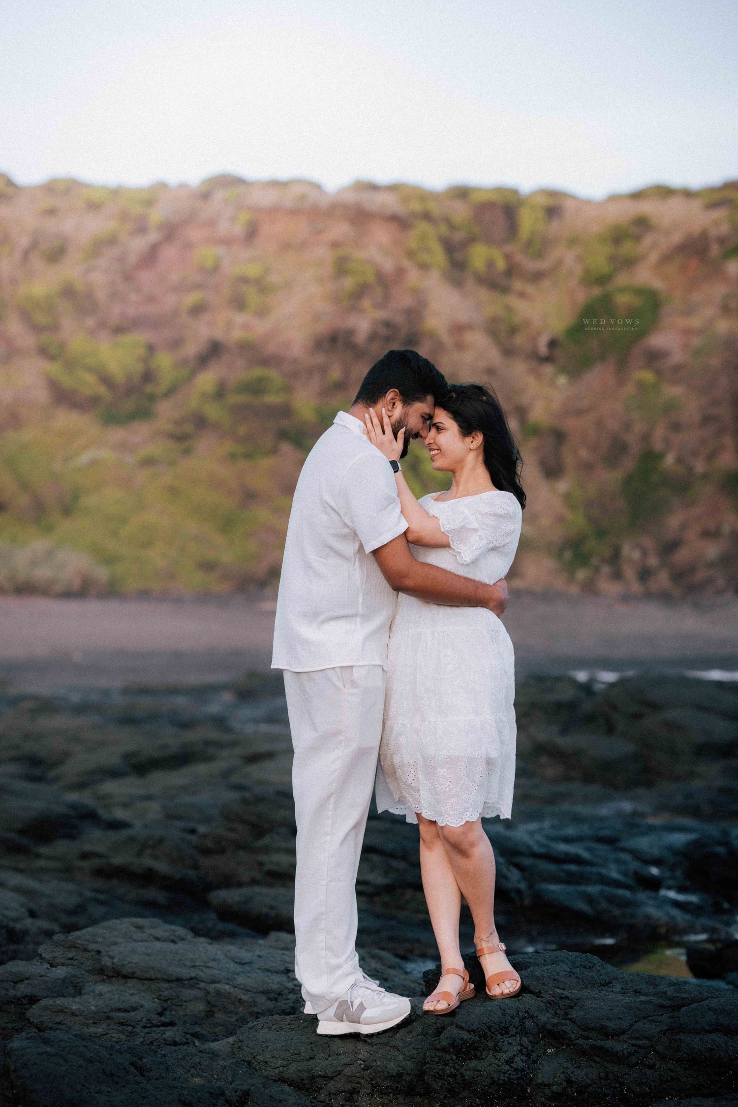 Couple in white touching foreheads intimately on rocky coastal terrain at dusk.