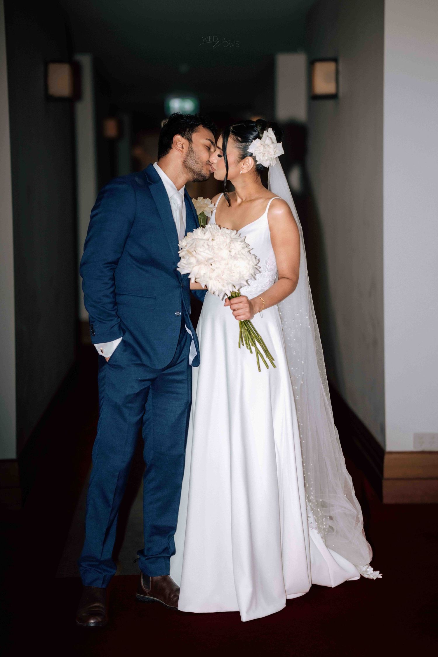 Bride and groom kissing in hotel corridor, bride in white gown holding white floral bouquet.