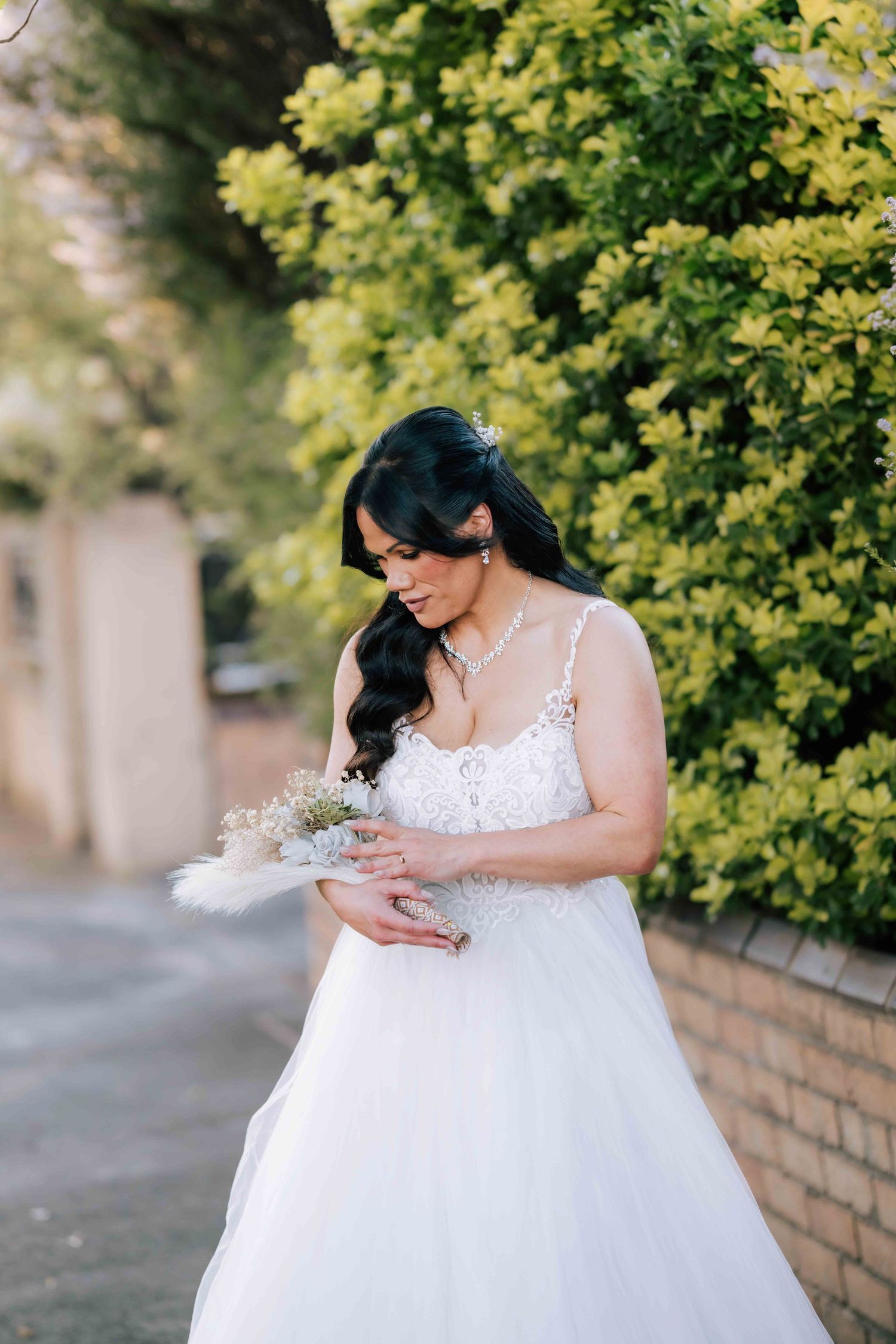 Dark-haired bride in a white lace wedding dress holding a bouquet outdoors near green hedges.