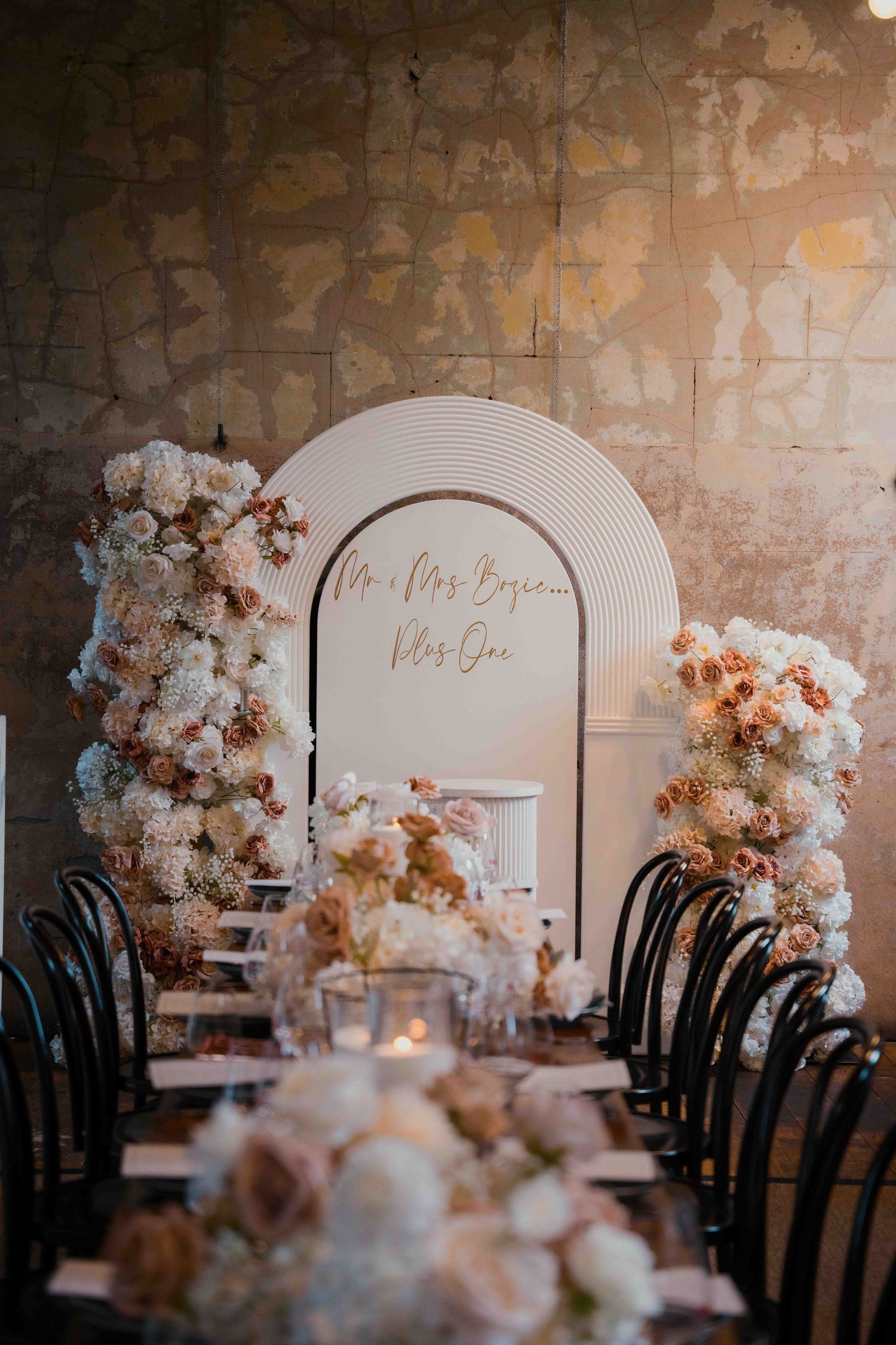 Wedding arch sign reading Mr and Mrs Bosie Plus One flanked by white and blush floral columns above reception table.
