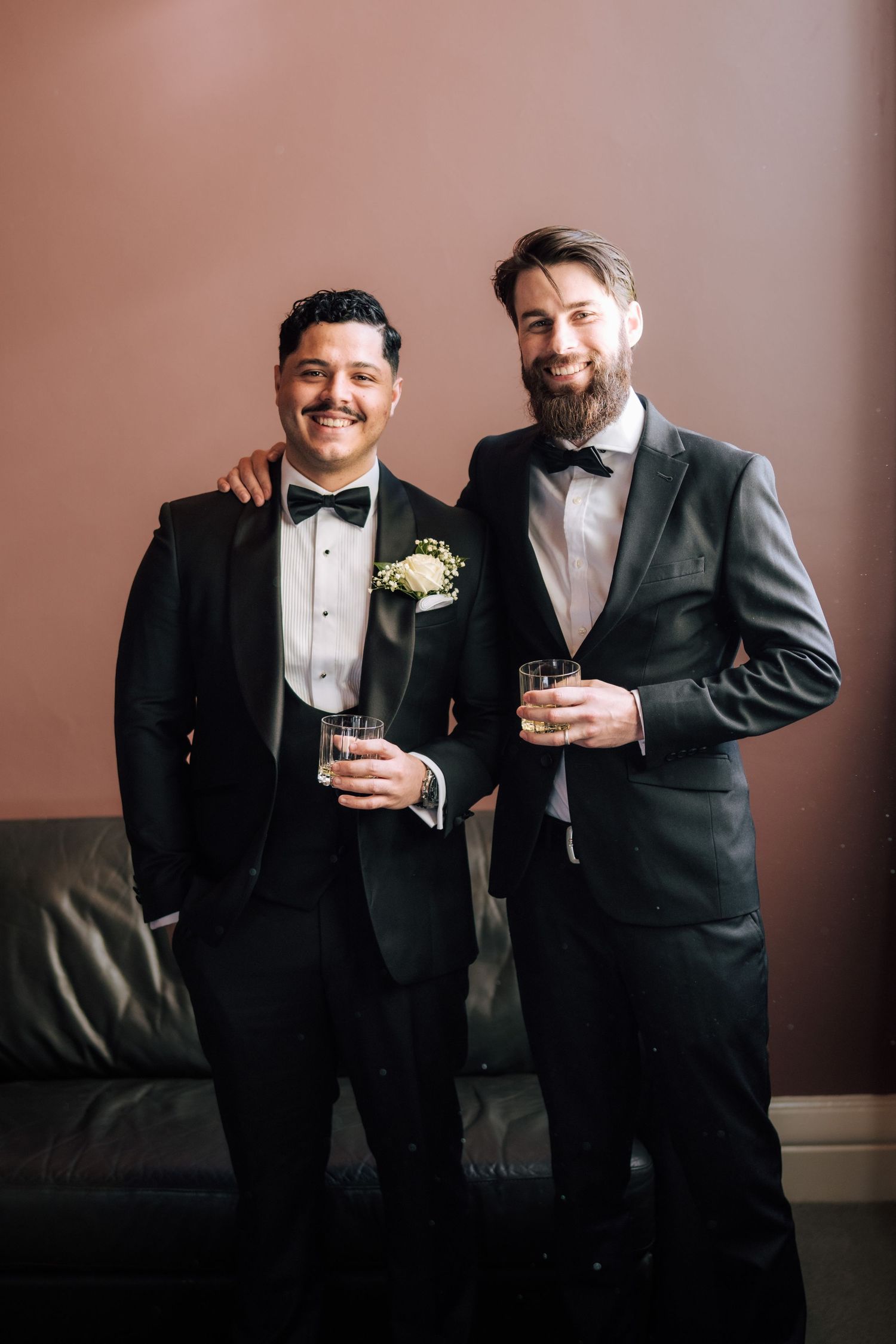 Two men in black tuxedos with bow ties holding drinks and smiling at wedding celebration.