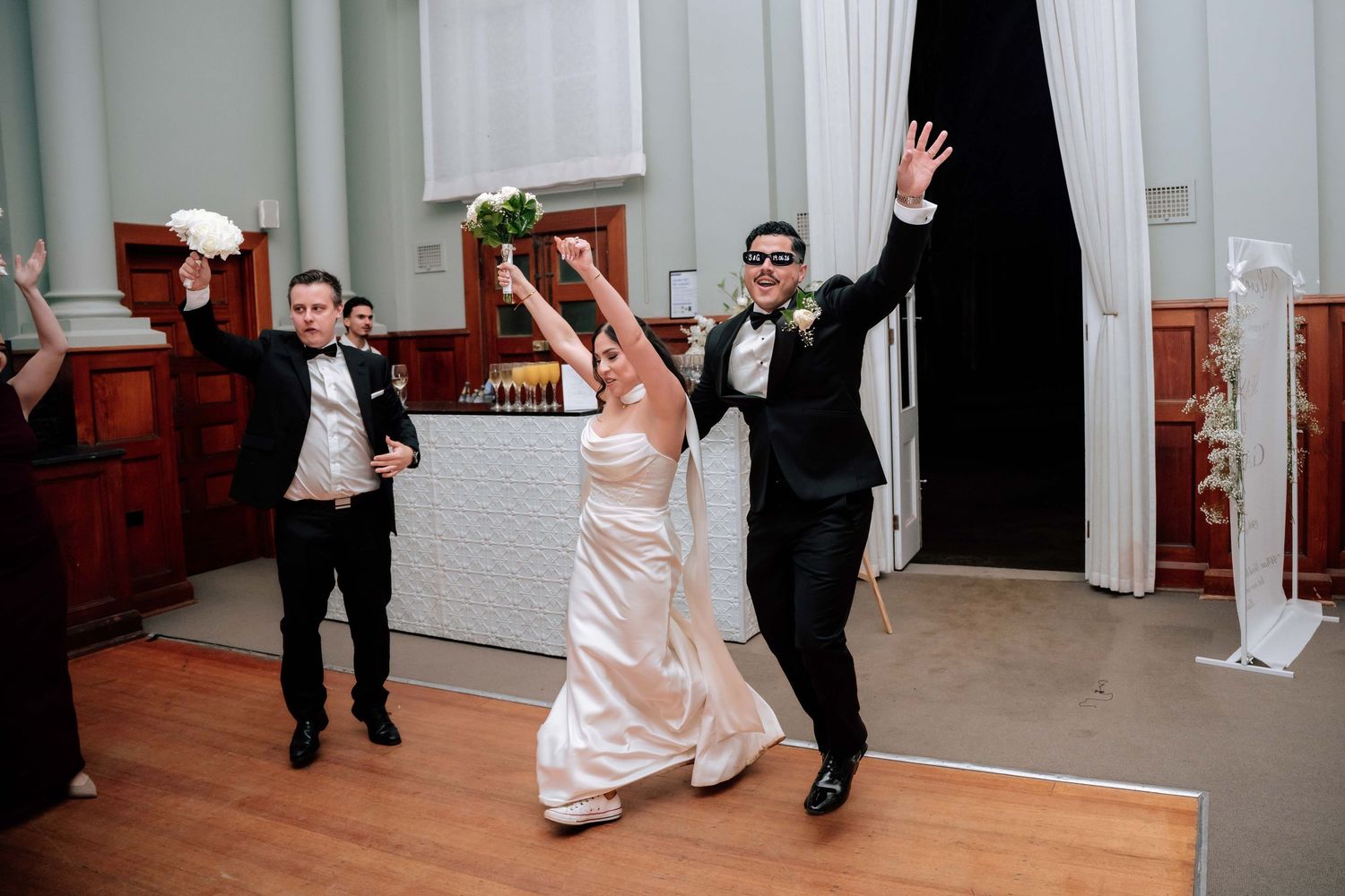 Bride and groom make a joyful entrance into wedding reception hall with raised arms and bouquet.