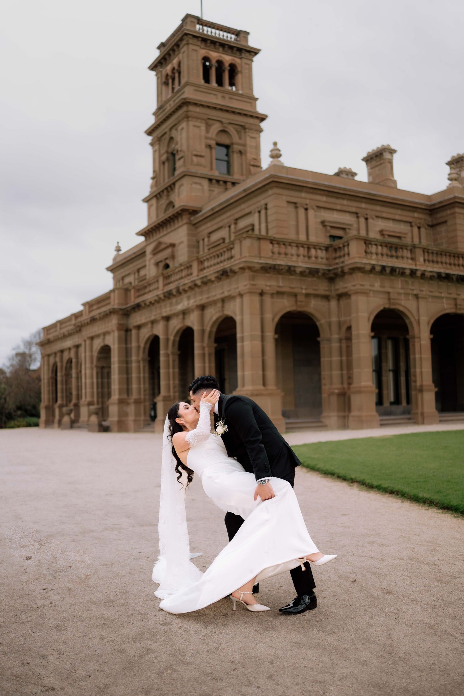 Groom dips bride in romantic kiss outside grand historic sandstone mansion on their wedding day.