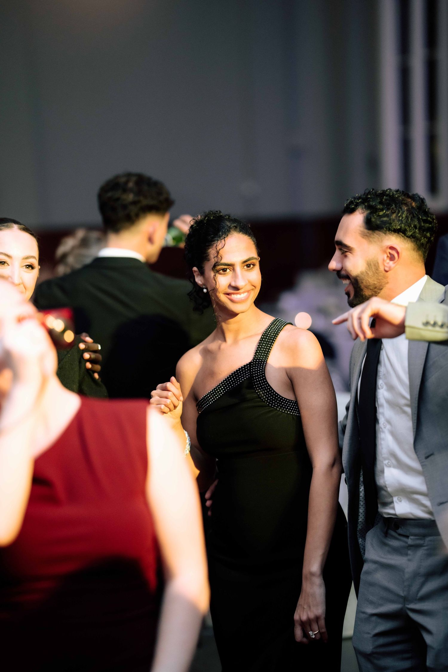 Woman in black one-shoulder dress smiling at a formal event while guests mingle in the background.