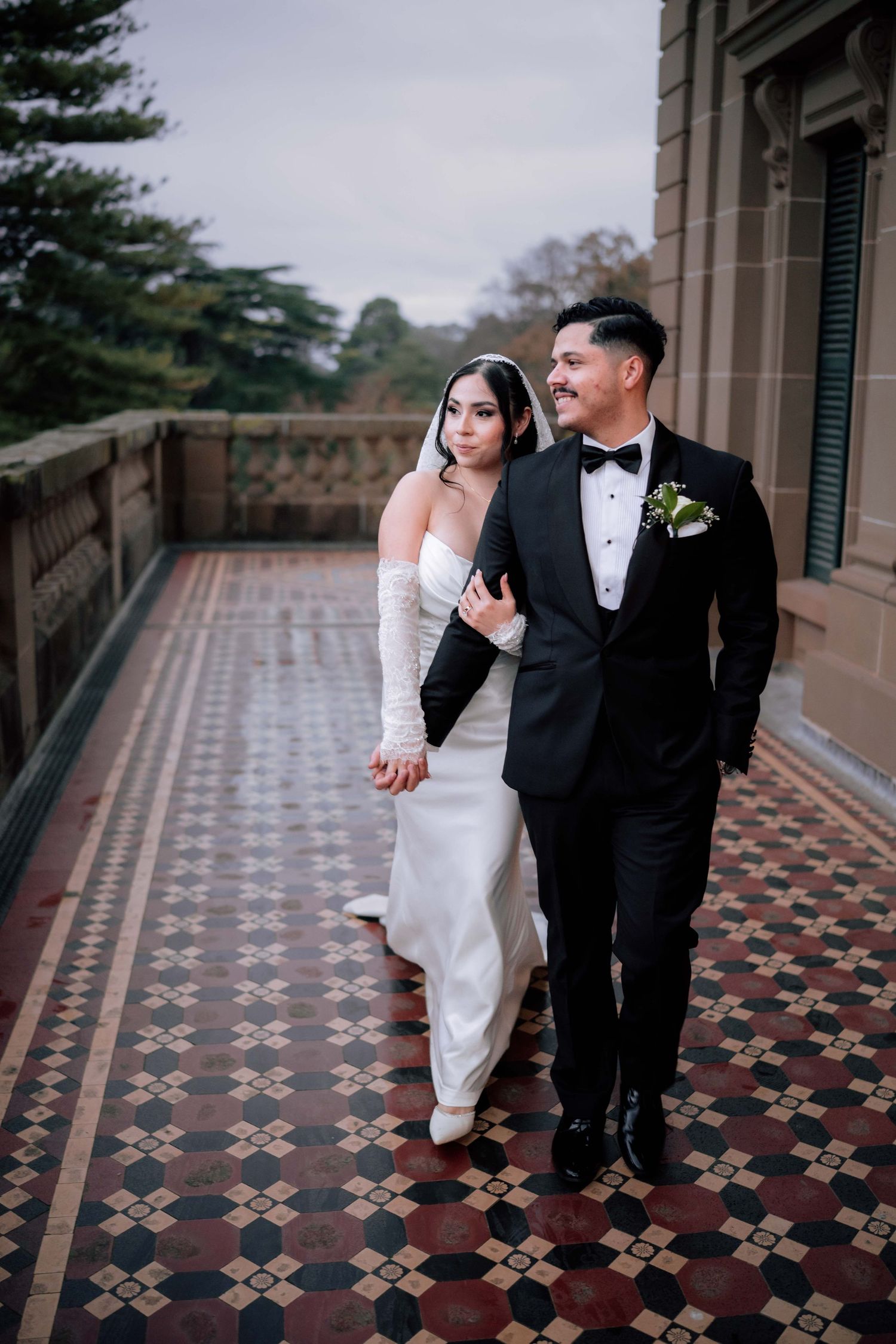 Bride and groom walking on ornate tiled balcony, she in white strapless gown, he in black tuxedo.