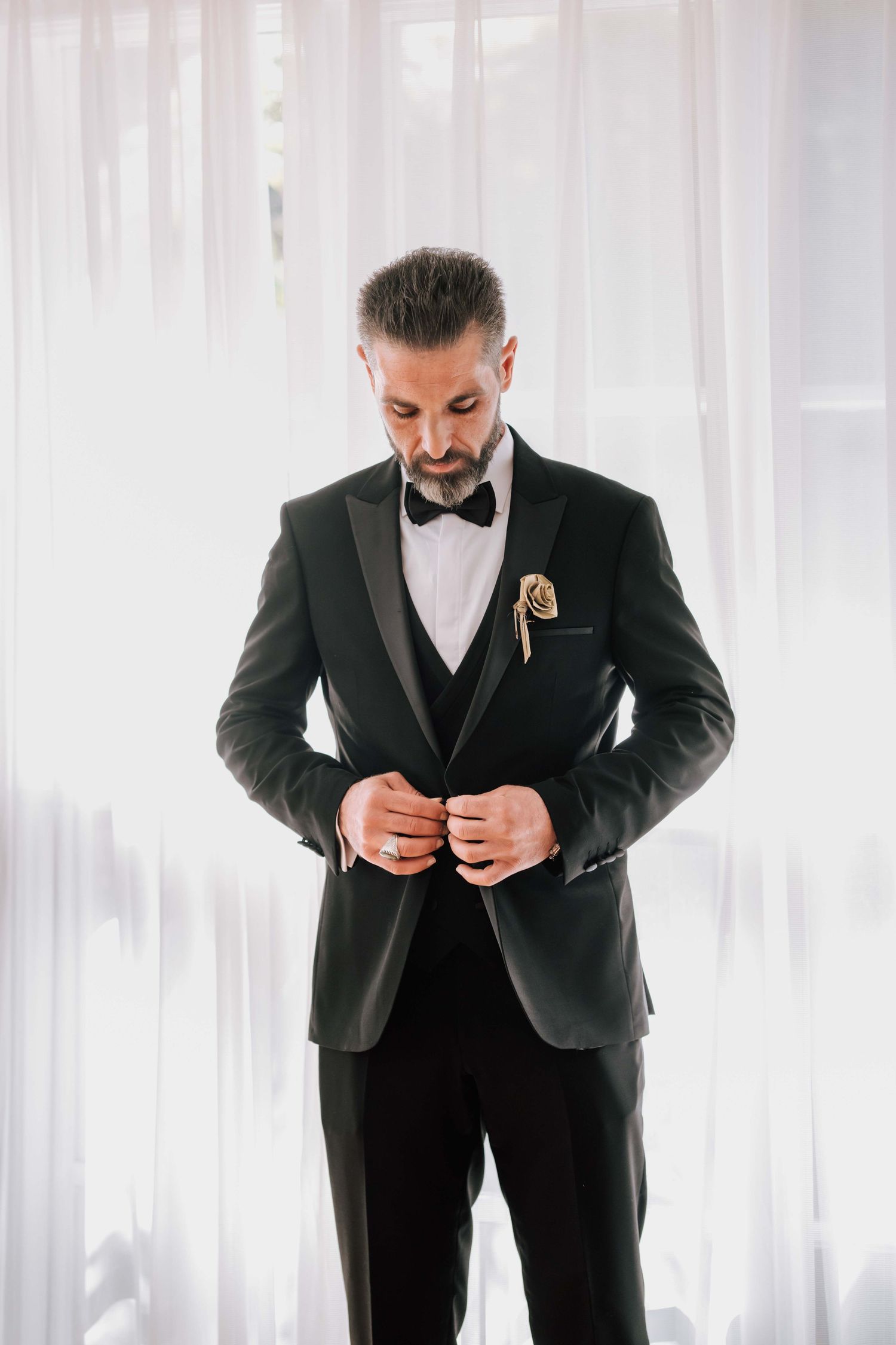 Dapper groom in black tuxedo with gold boutonniere buttoning jacket while standing by sheer curtains.