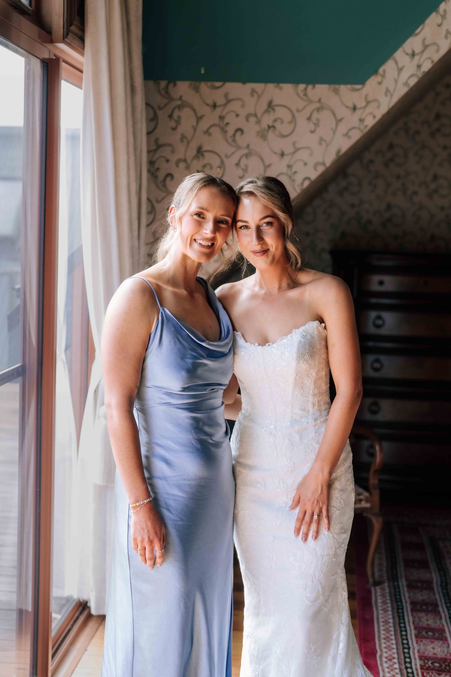 Bride in white lace gown poses with bridesmaid in blue satin dress near sunlit window.