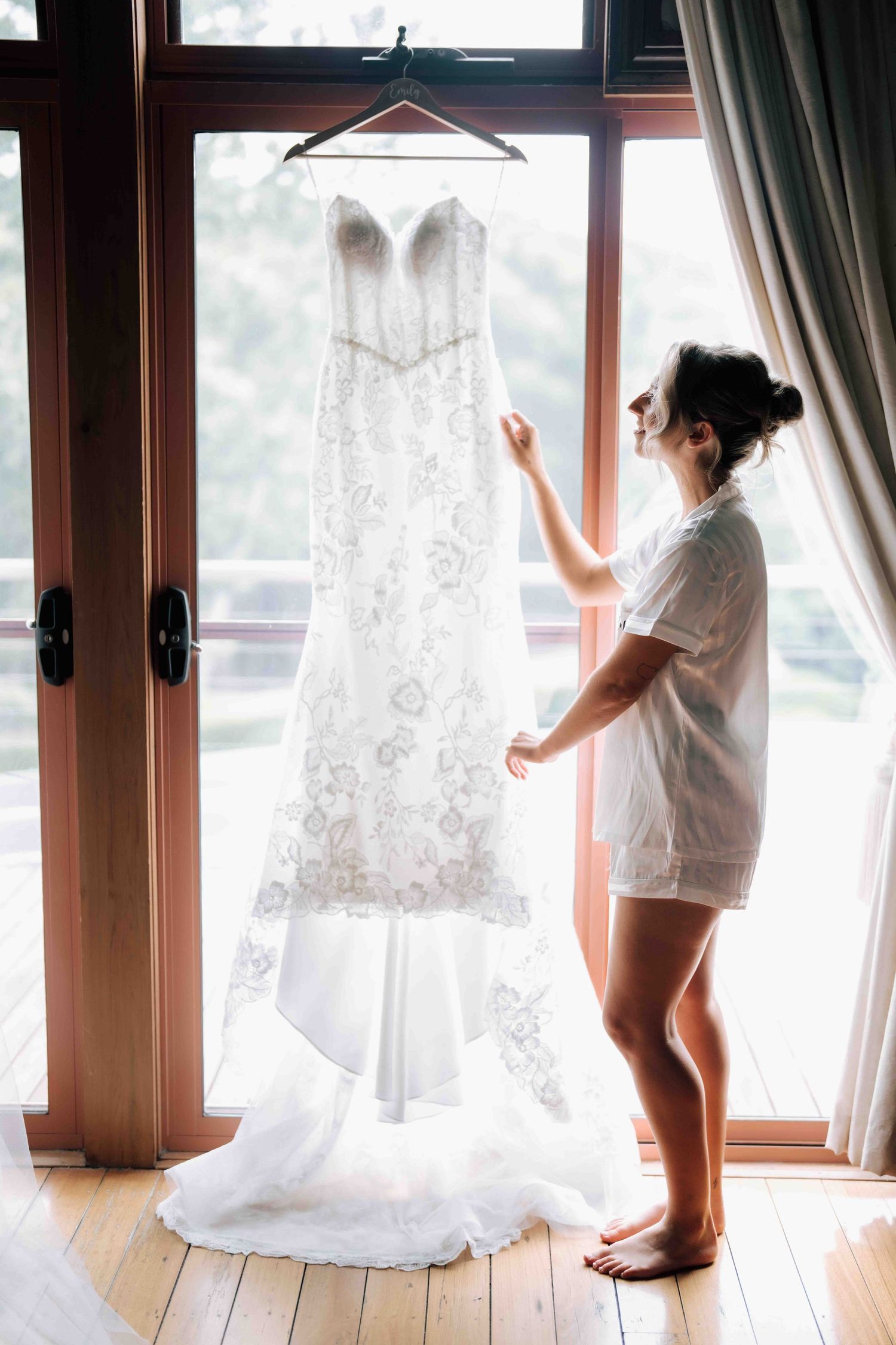 Bride in white pajamas admiring her lace wedding dress hanging by a window.