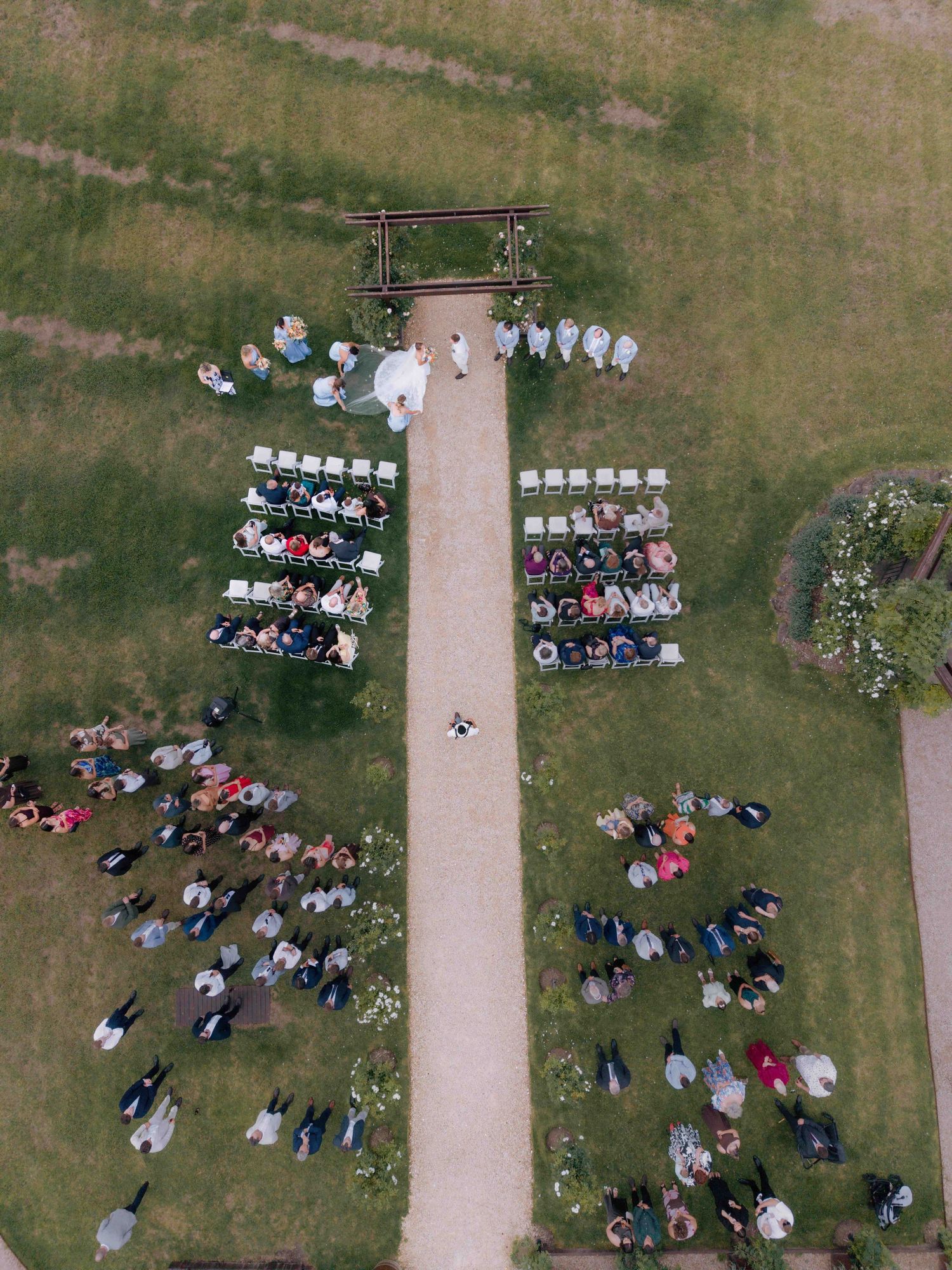 Aerial drone view of outdoor wedding ceremony with guests seated and standing around a garden aisle.