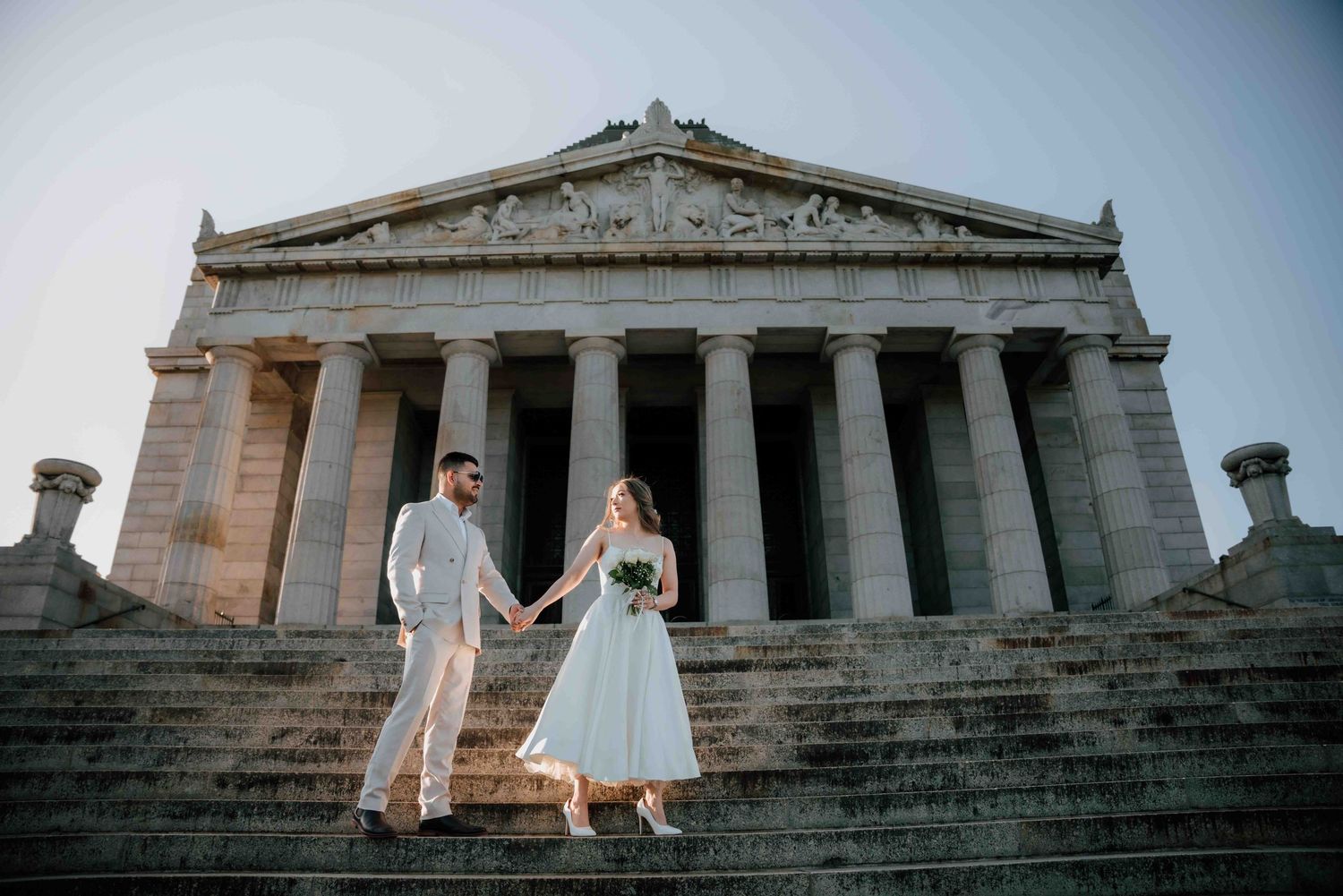 Bride and groom hold hands on steps of neoclassical building with grand columns during wedding photoshoot.