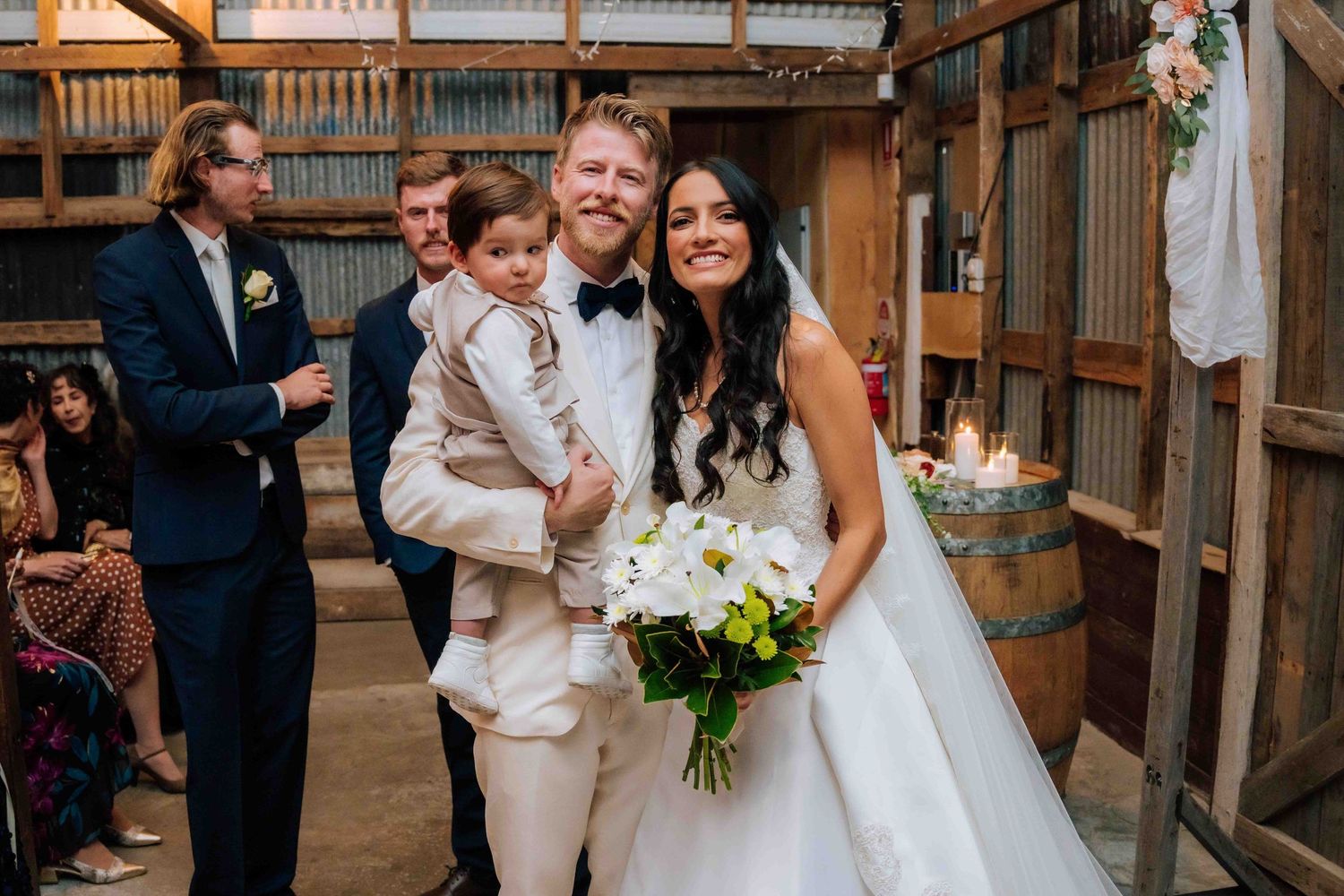 Bride in white gown holding bouquet stands with groom in tuxedo holding young boy at rustic barn wedding ceremony.
