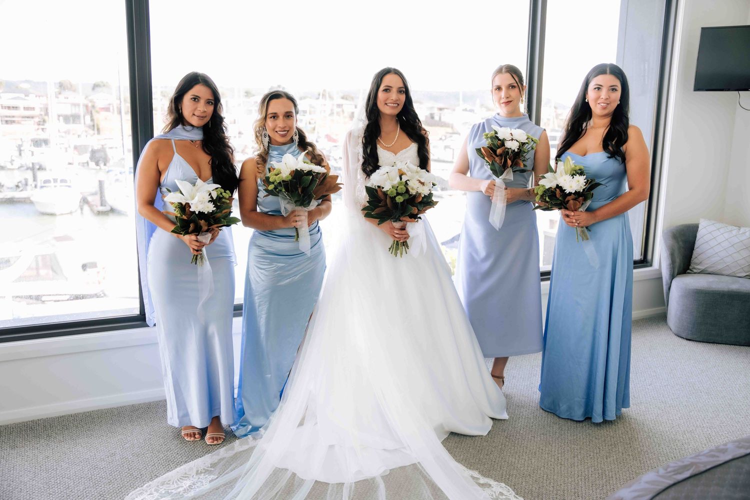 Bride in white ball gown poses with four bridesmaids in light blue satin dresses holding bouquets indoors.