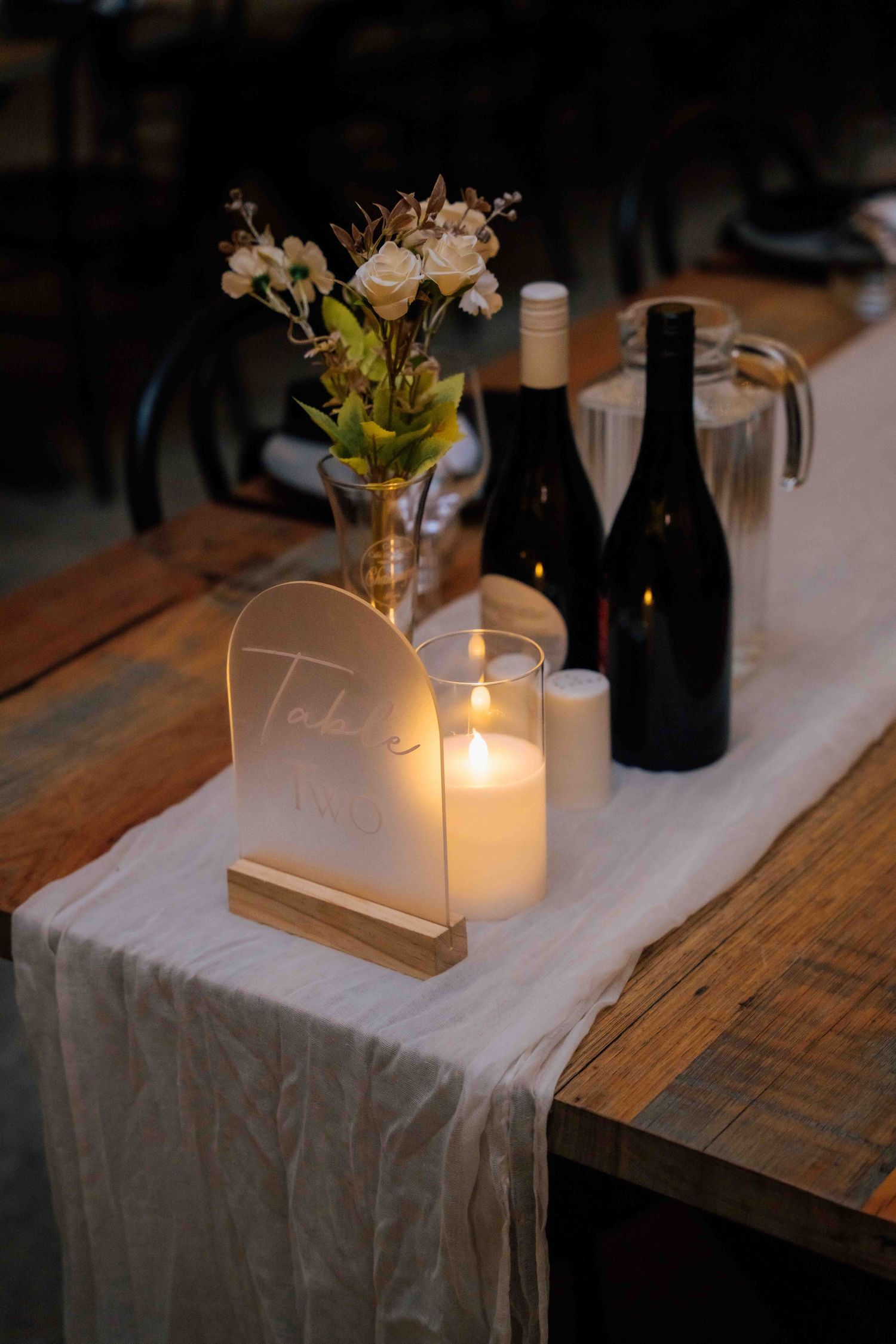 Elegant wedding table centerpiece with candle, white roses, table number sign and wine bottles.