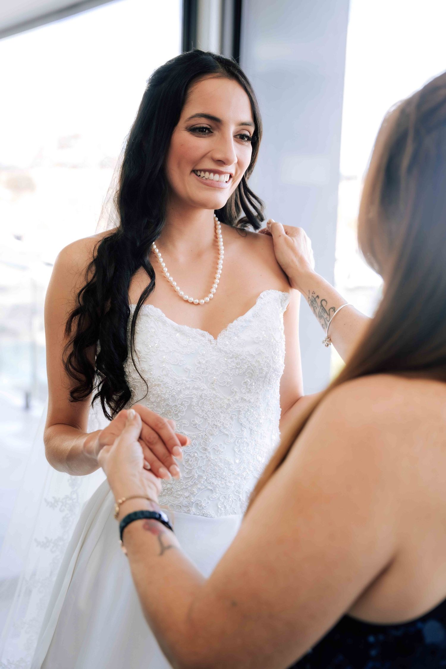 Smiling dark-haired bride in lace wedding dress and pearl necklace being helped by bridesmaid near bright window.
